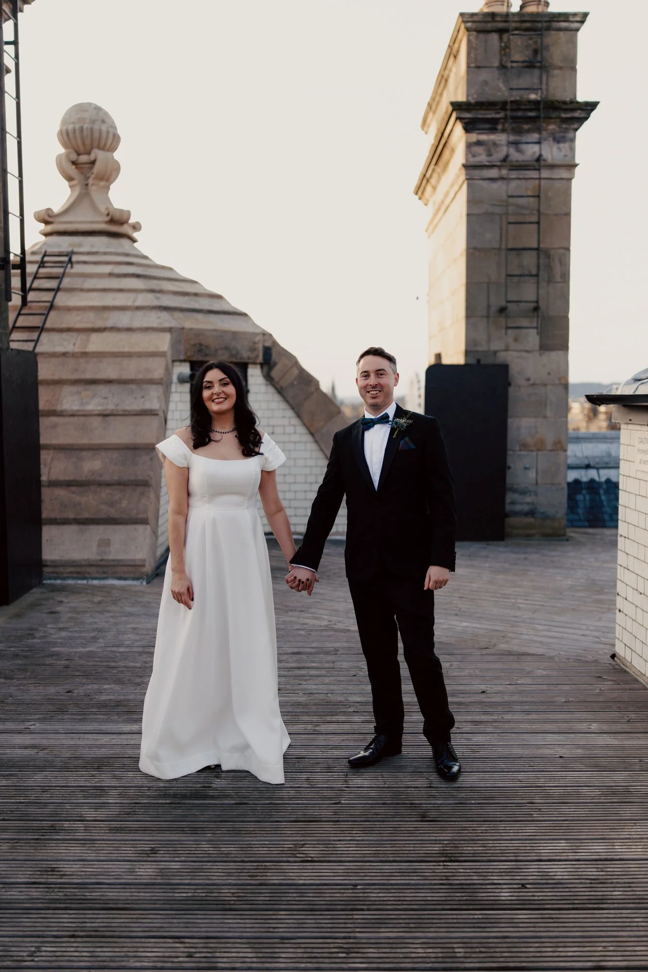 Full-length rooftop portrait of the couple beside a stone chimney.