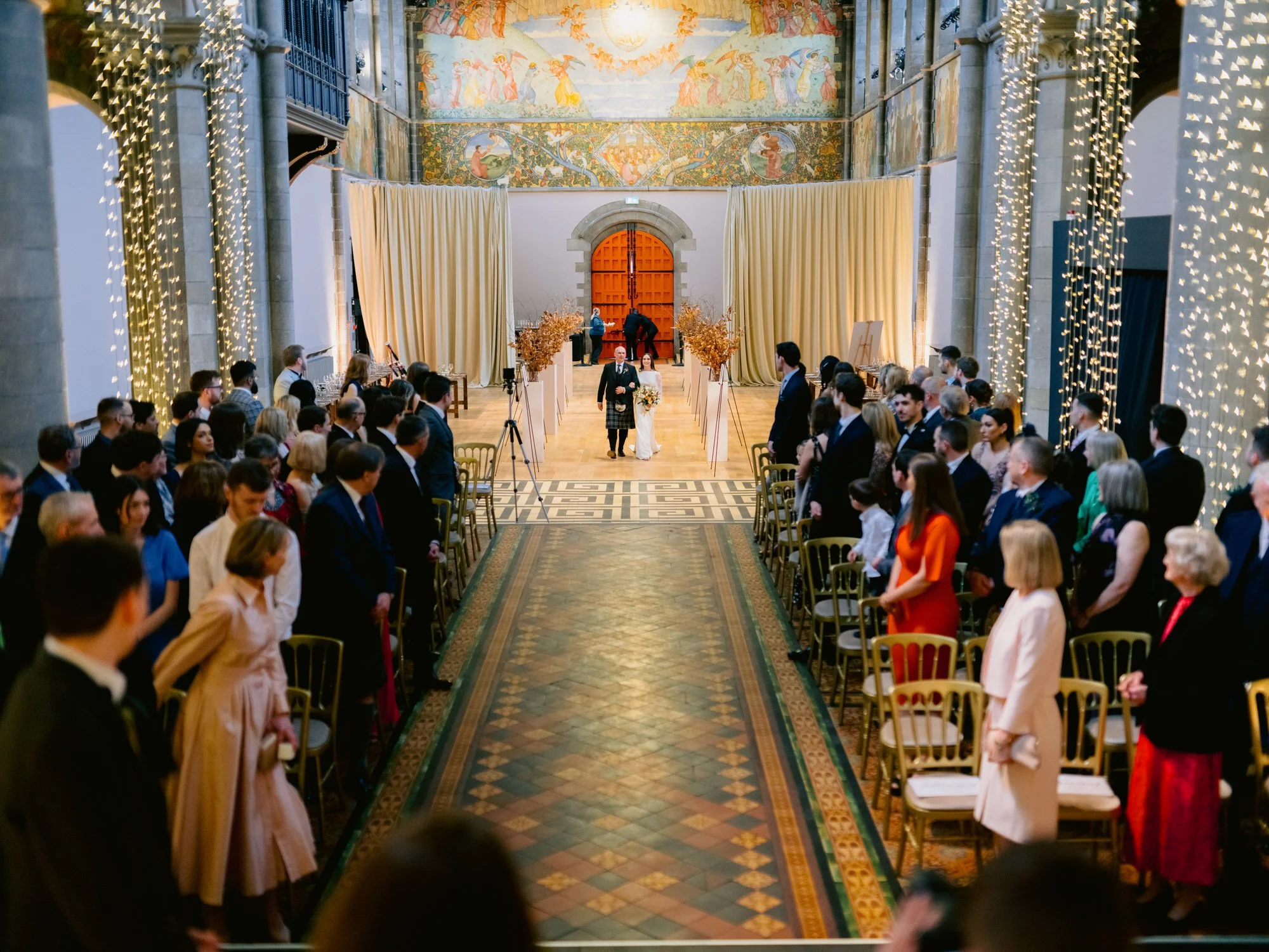 Nicola entering the tiled aisle between seated wedding guests