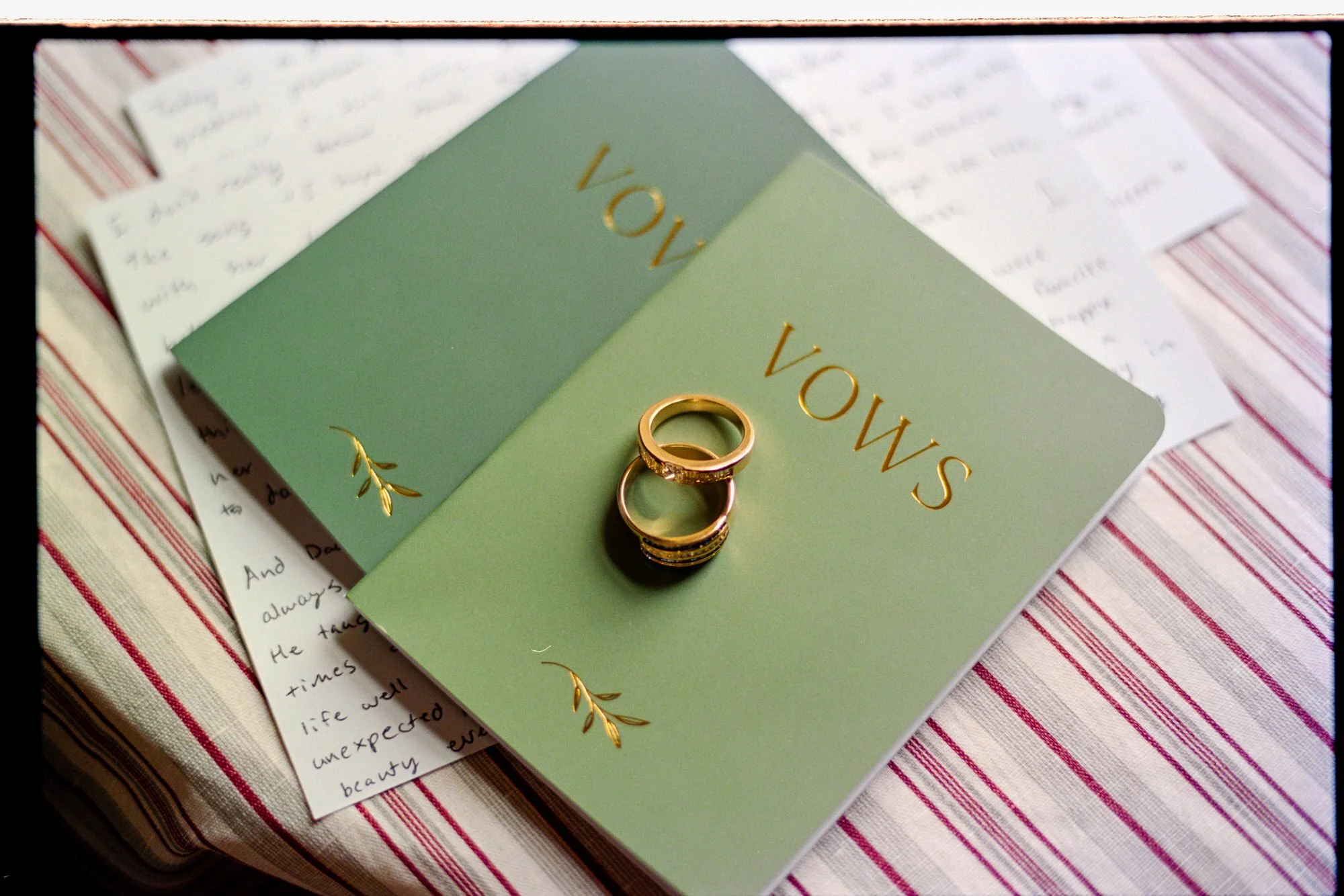 Two gold wedding rings resting on green vow books and handwritten notes.