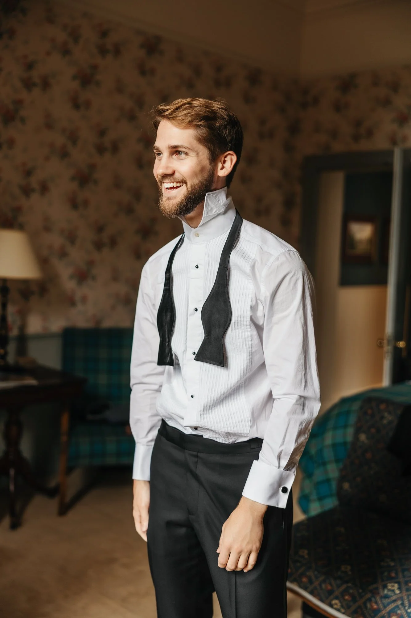A groom smiling in a white shirt with his untied bow tie around his neck.