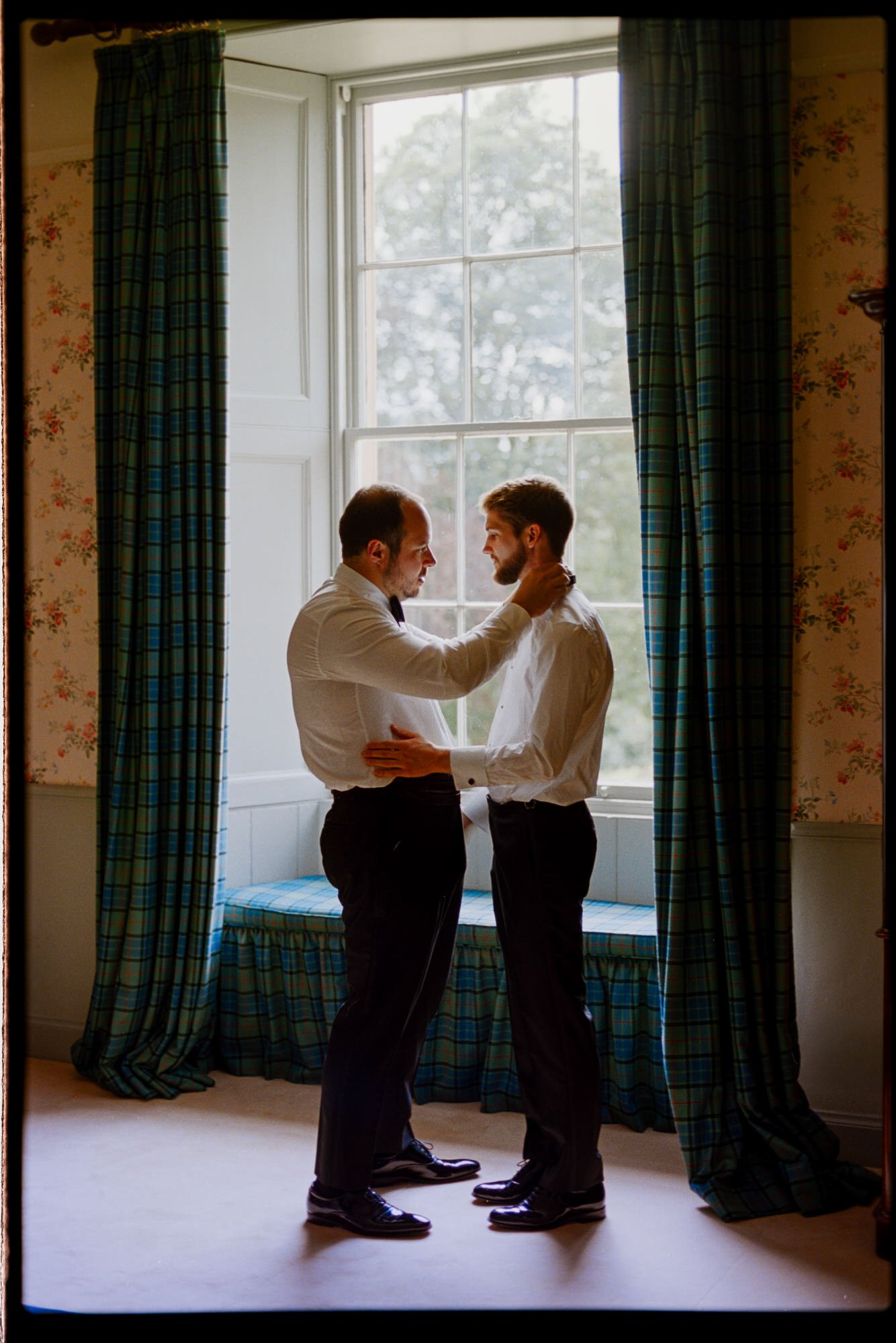 The grooms getting ready together beside a tall window with tartan curtains.
