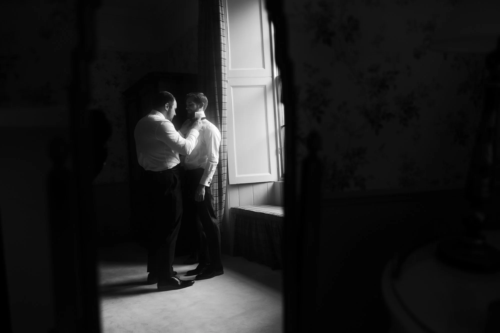 Black-and-white photograph of the grooms adjusting a bow tie in window light.