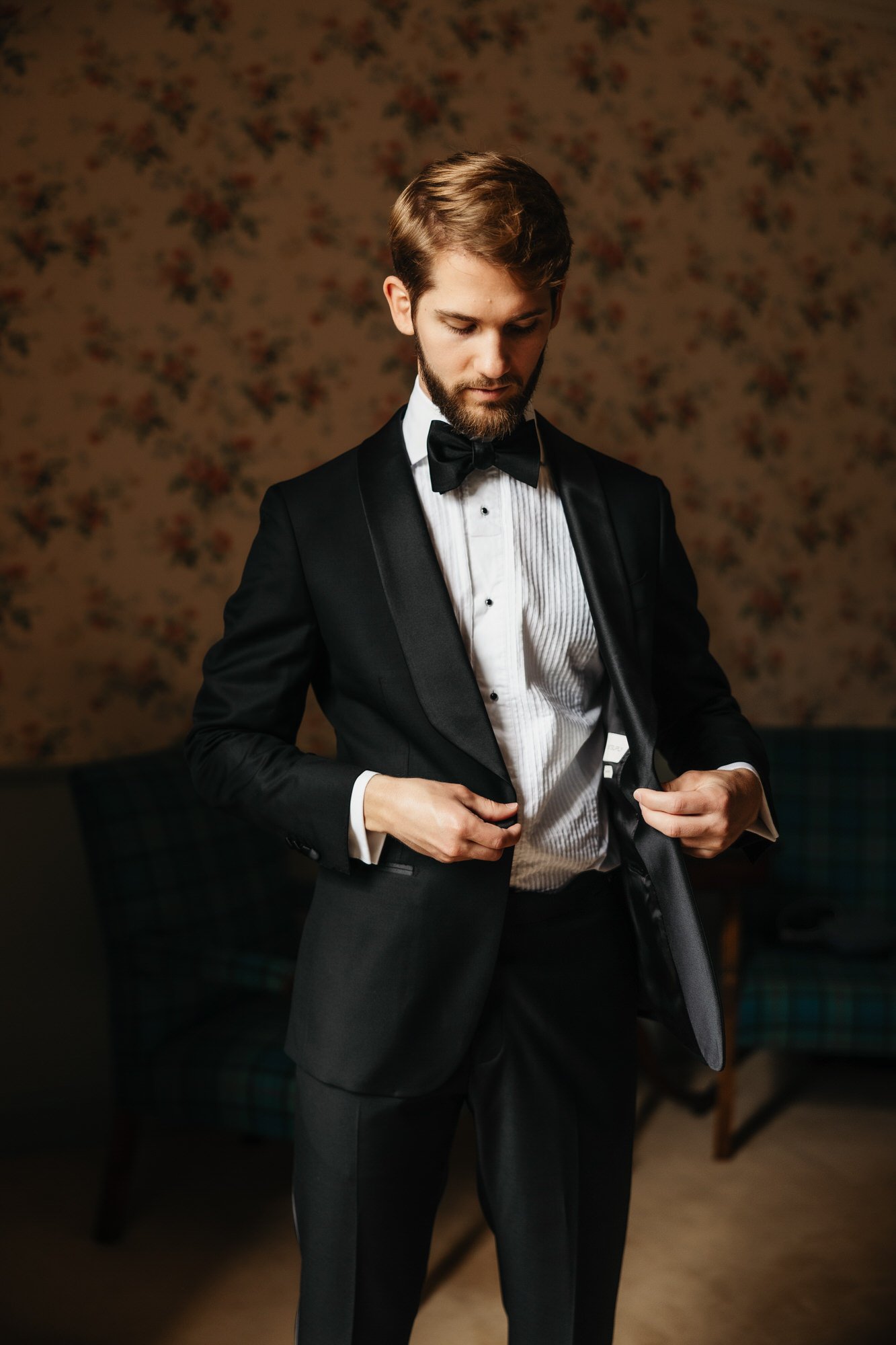 A groom adjusting his dinner jacket in a floral-wallpapered room.