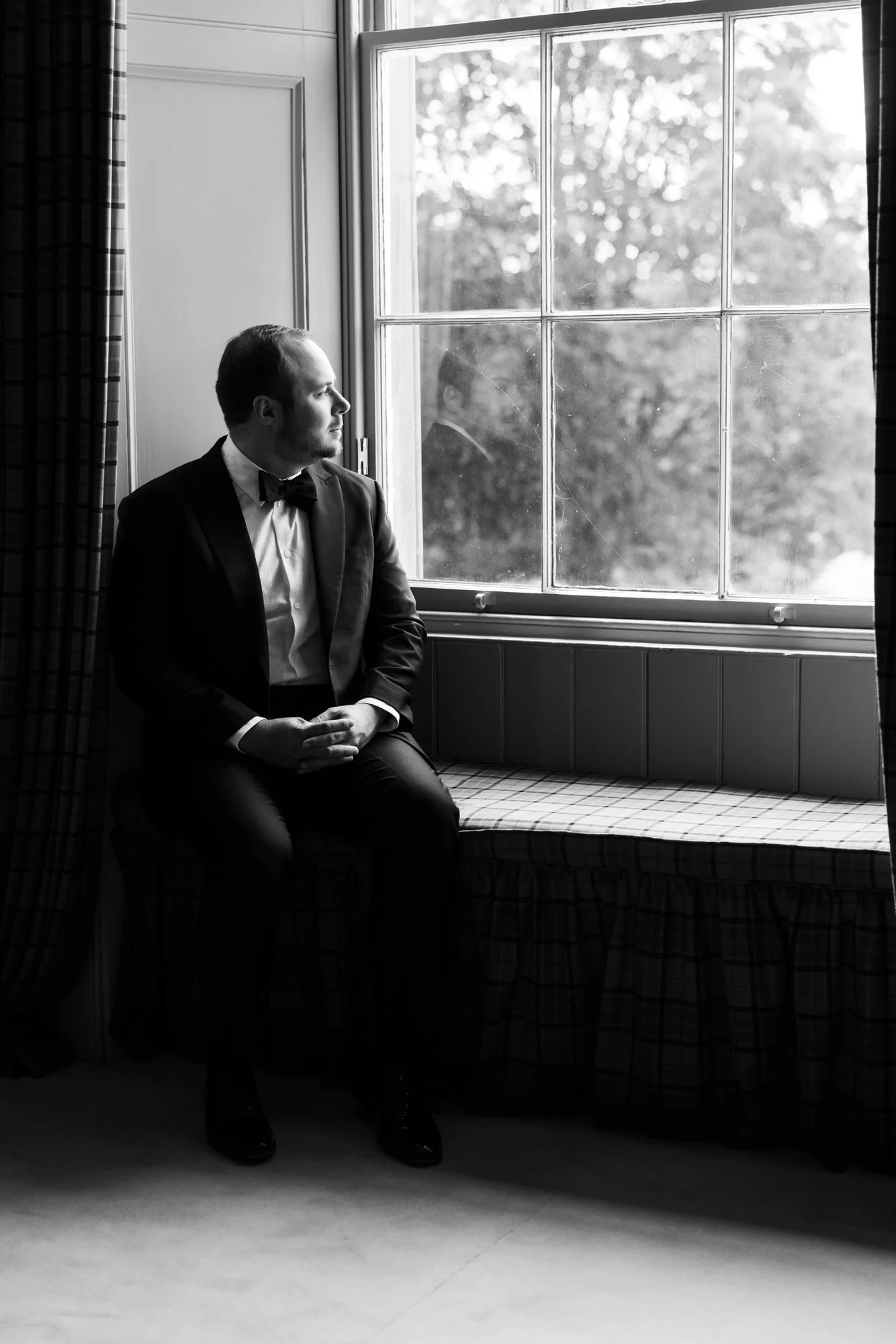 Black-and-white portrait of a groom sitting beside a tall window.