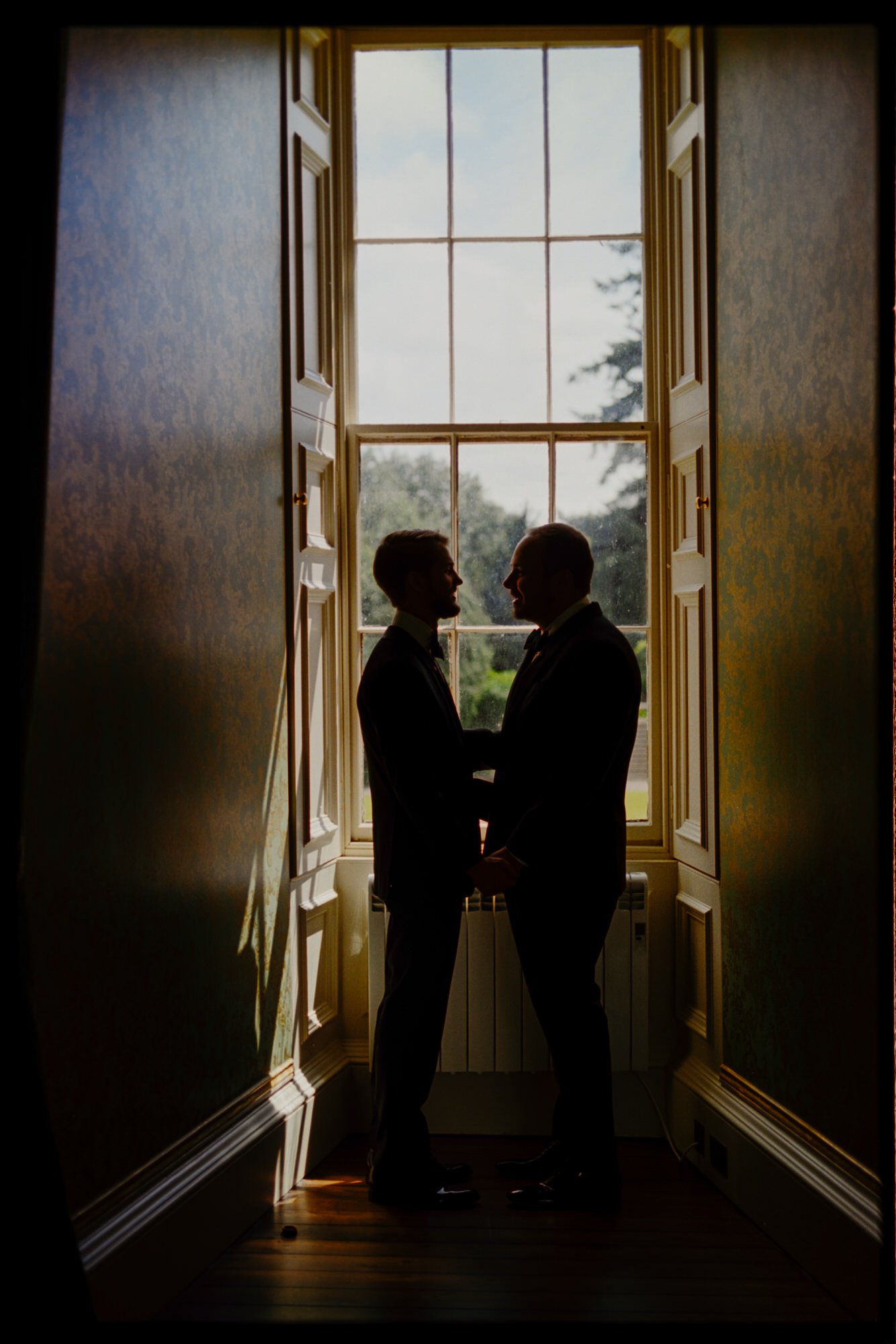 The grooms facing each other in silhouette beside a sunlit window.