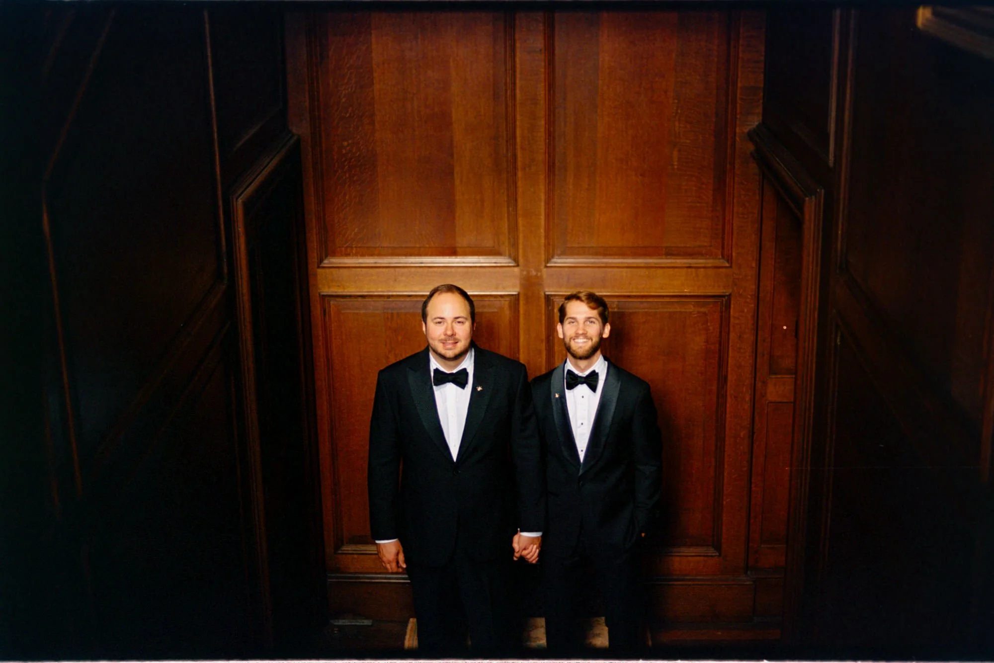 The grooms holding hands in front of a wooden panelled doorway.