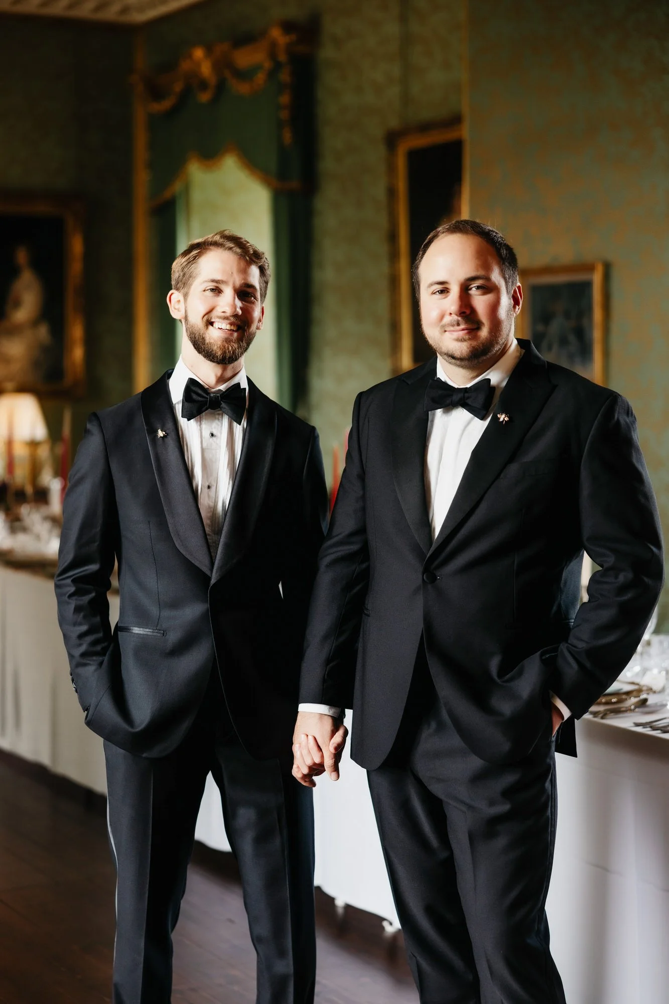 The grooms holding hands beneath portraits in an ornate castle room.
