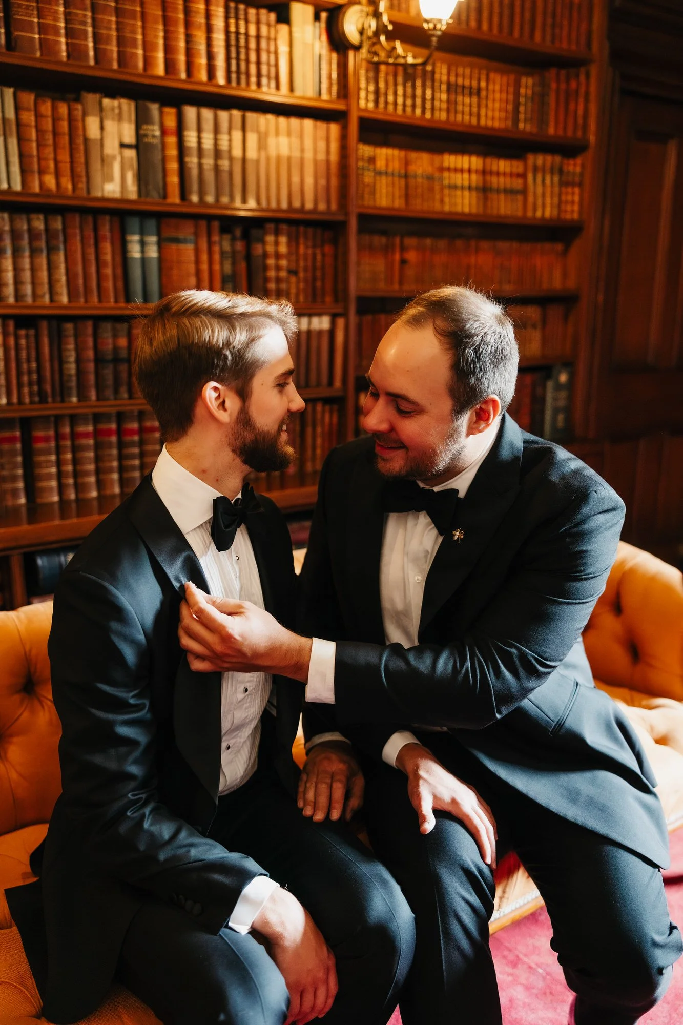 One groom playfully adjusting the other's bow tie on a library sofa.