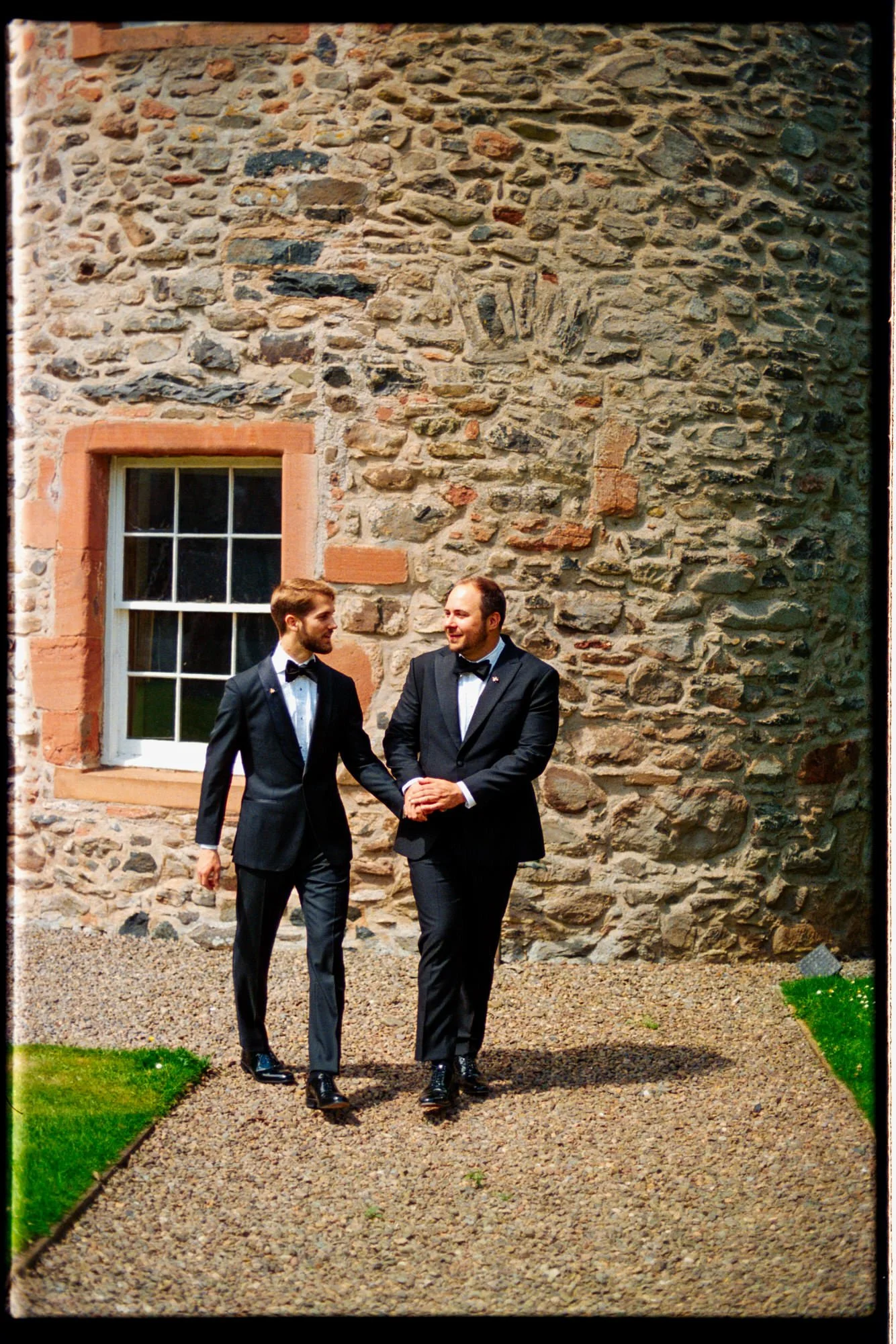 The grooms standing together on a gravel path beside the castle's stone walls.
