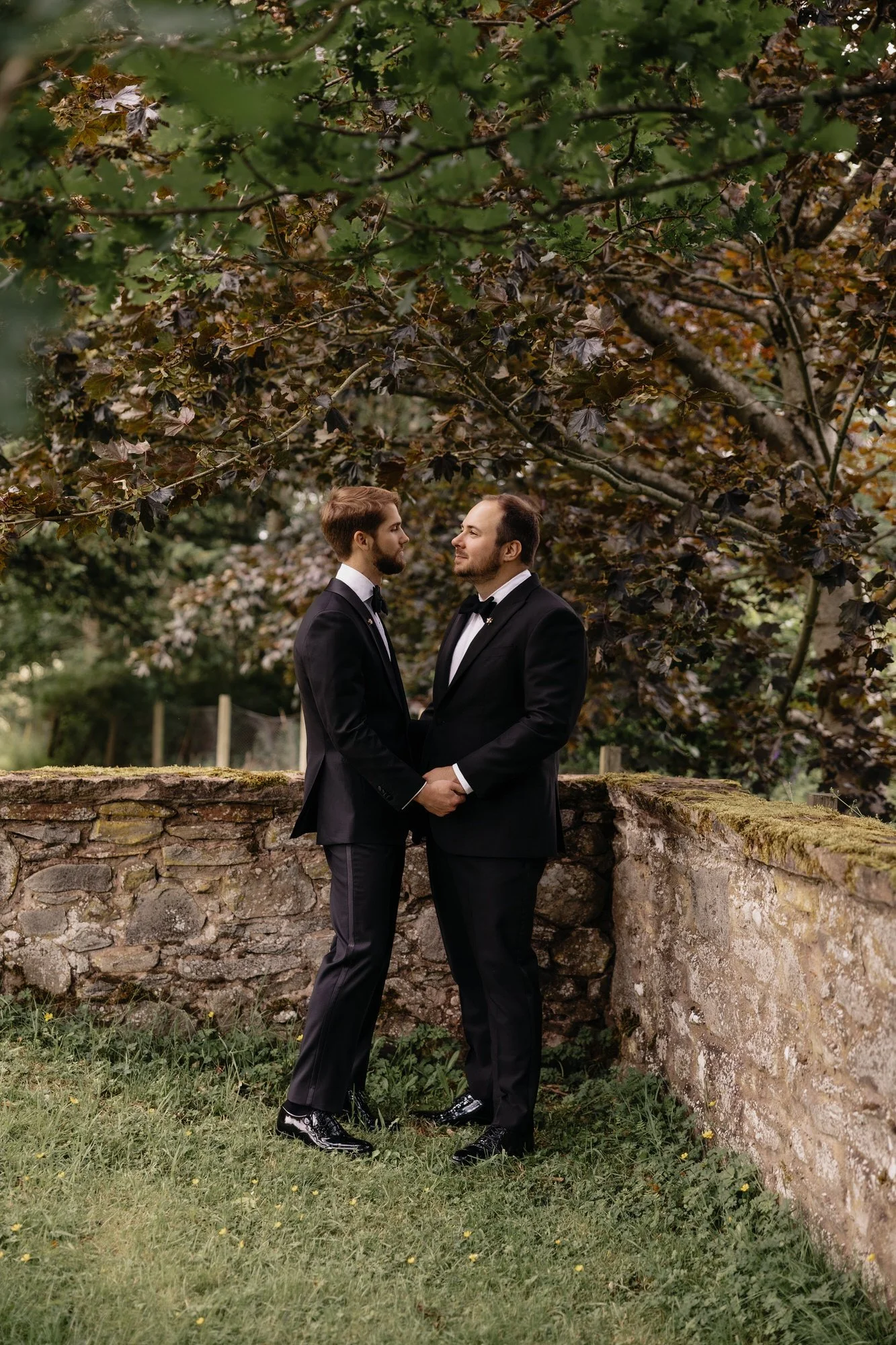 The grooms standing beside a low stone wall under the trees.