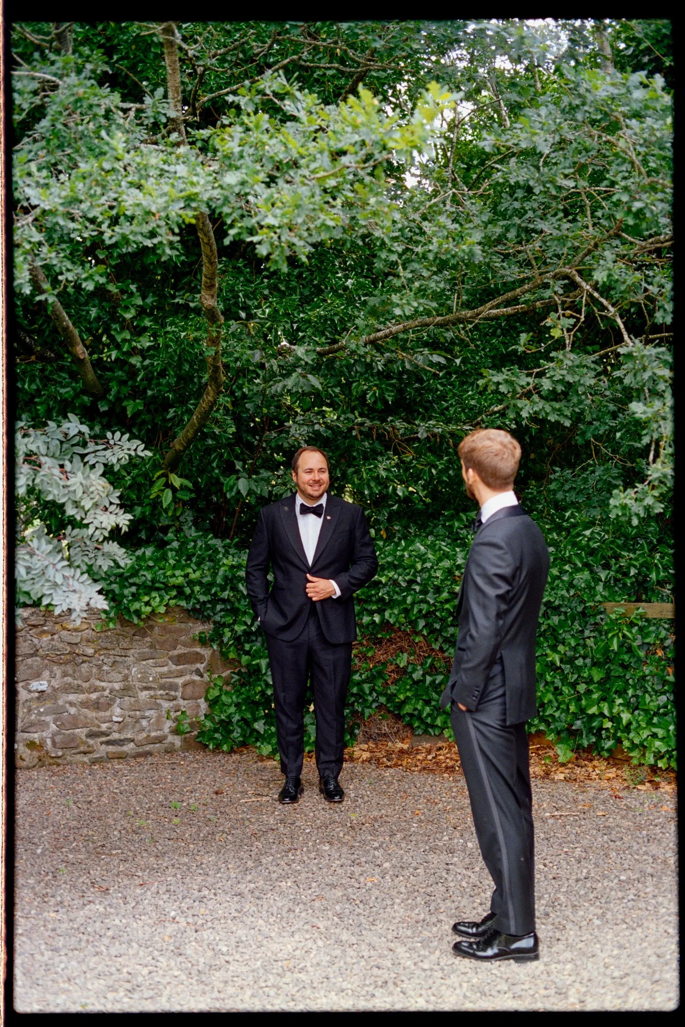 The grooms facing each other on a gravel path surrounded by greenery.