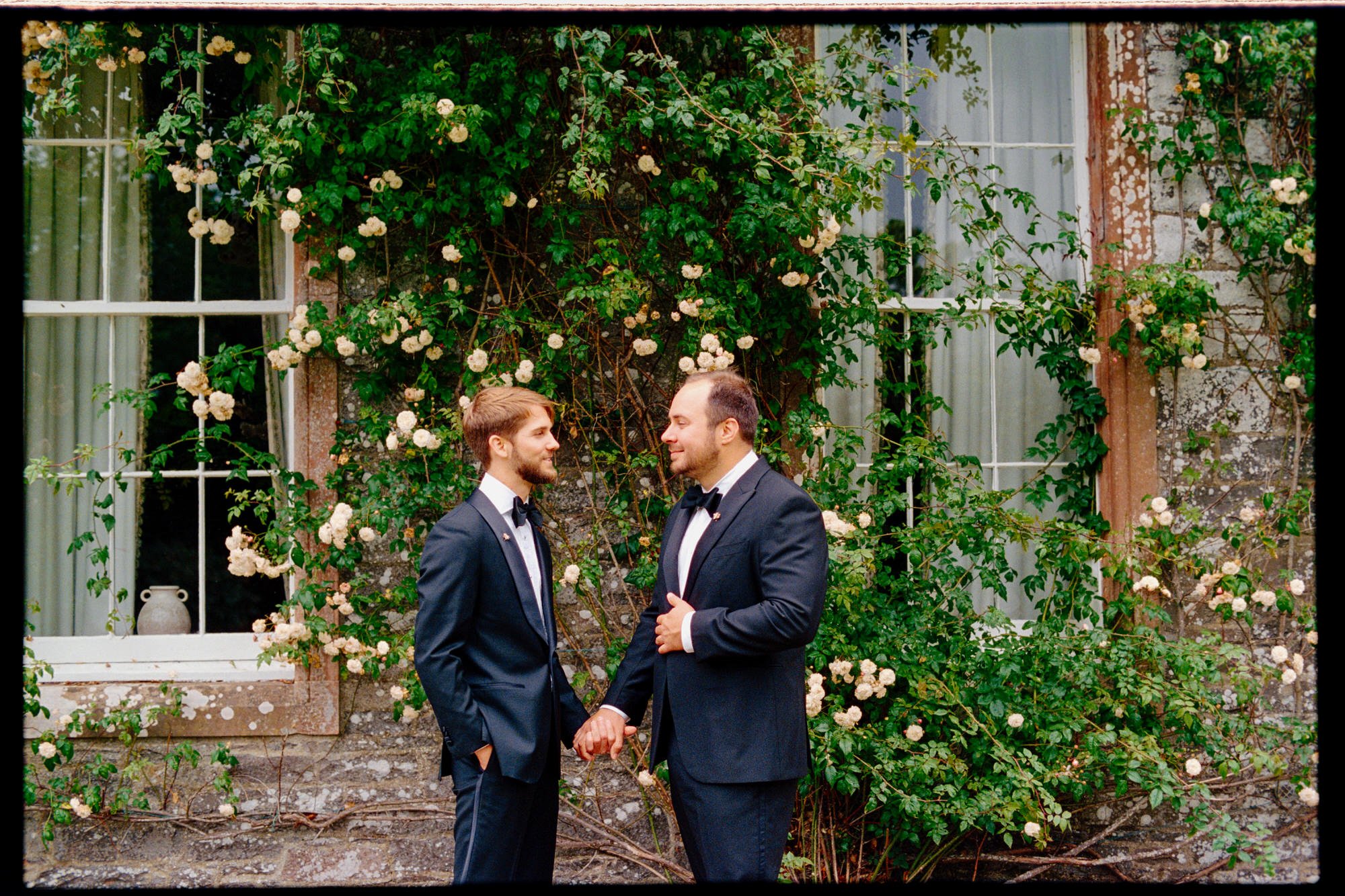 The grooms holding hands beside climbing pink roses and a castle window.
