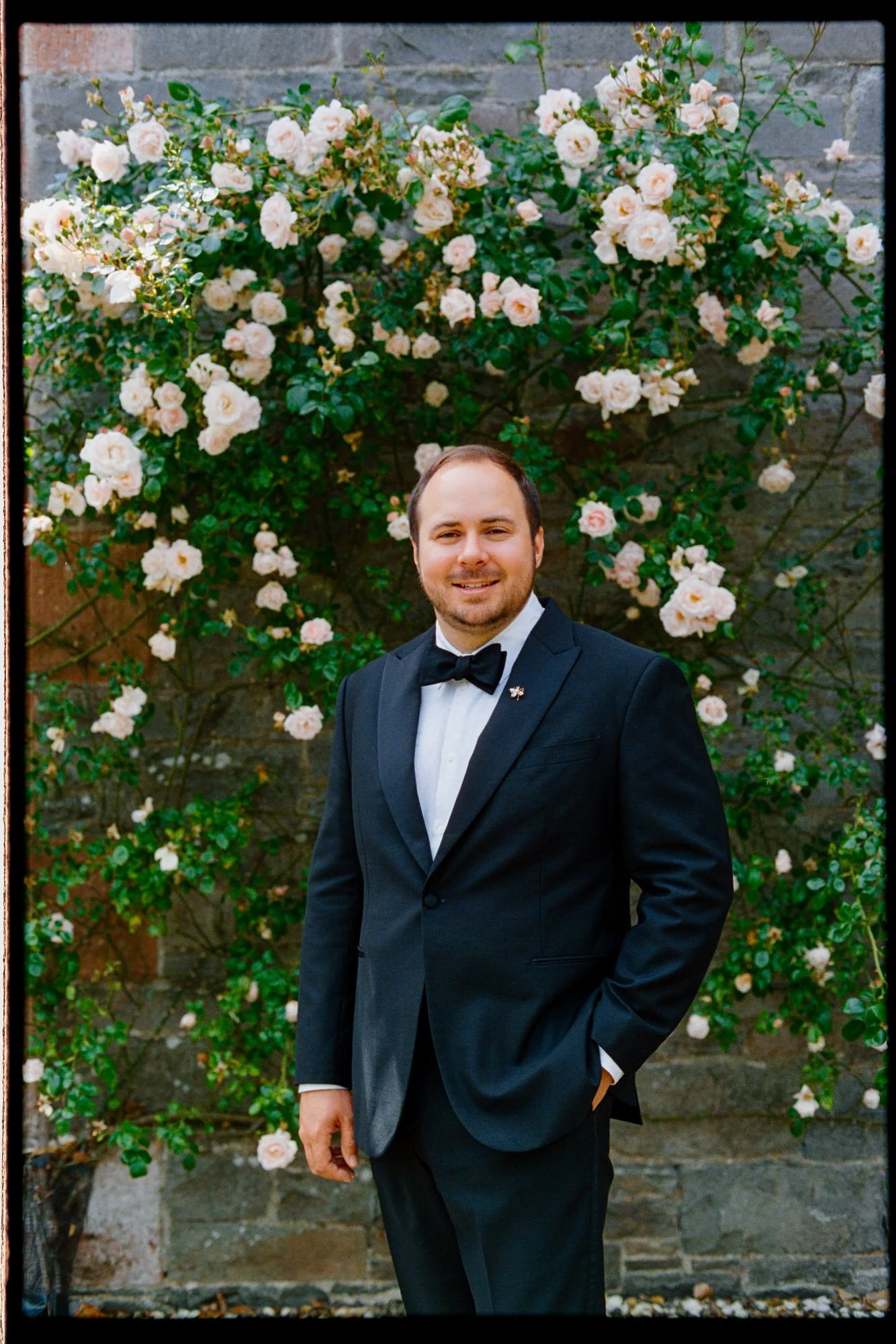 A groom smiling beside pink roses growing against the castle wall.