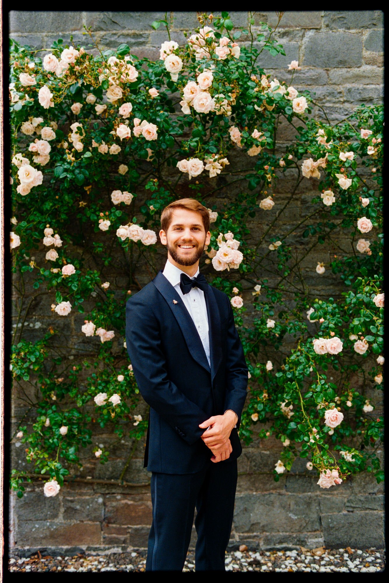 A groom in a dark dinner jacket standing beside climbing roses.