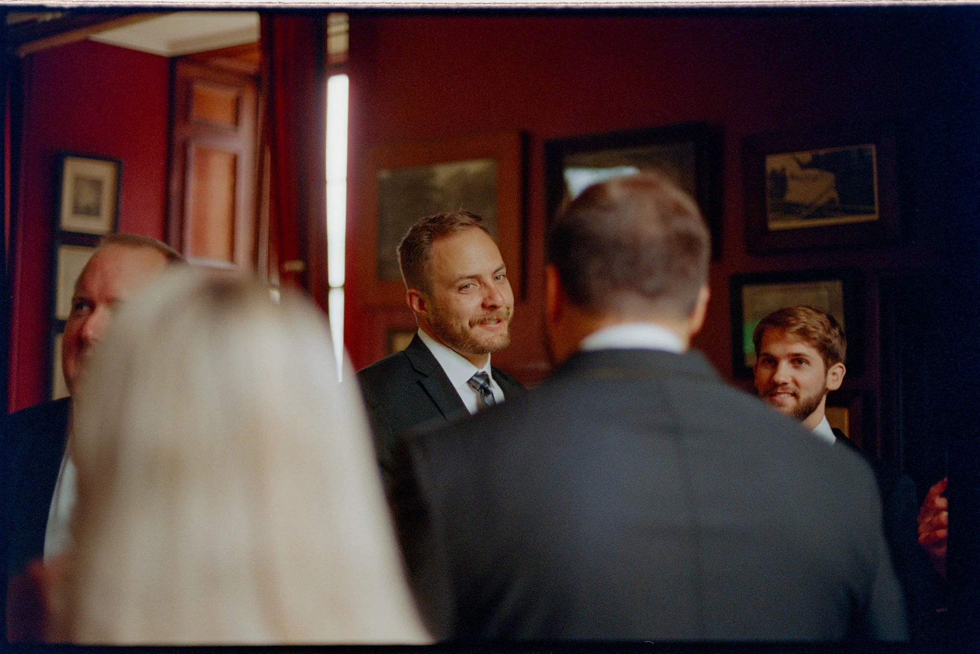 A groom smiling during a conversation with guests in a wood-panelled room.