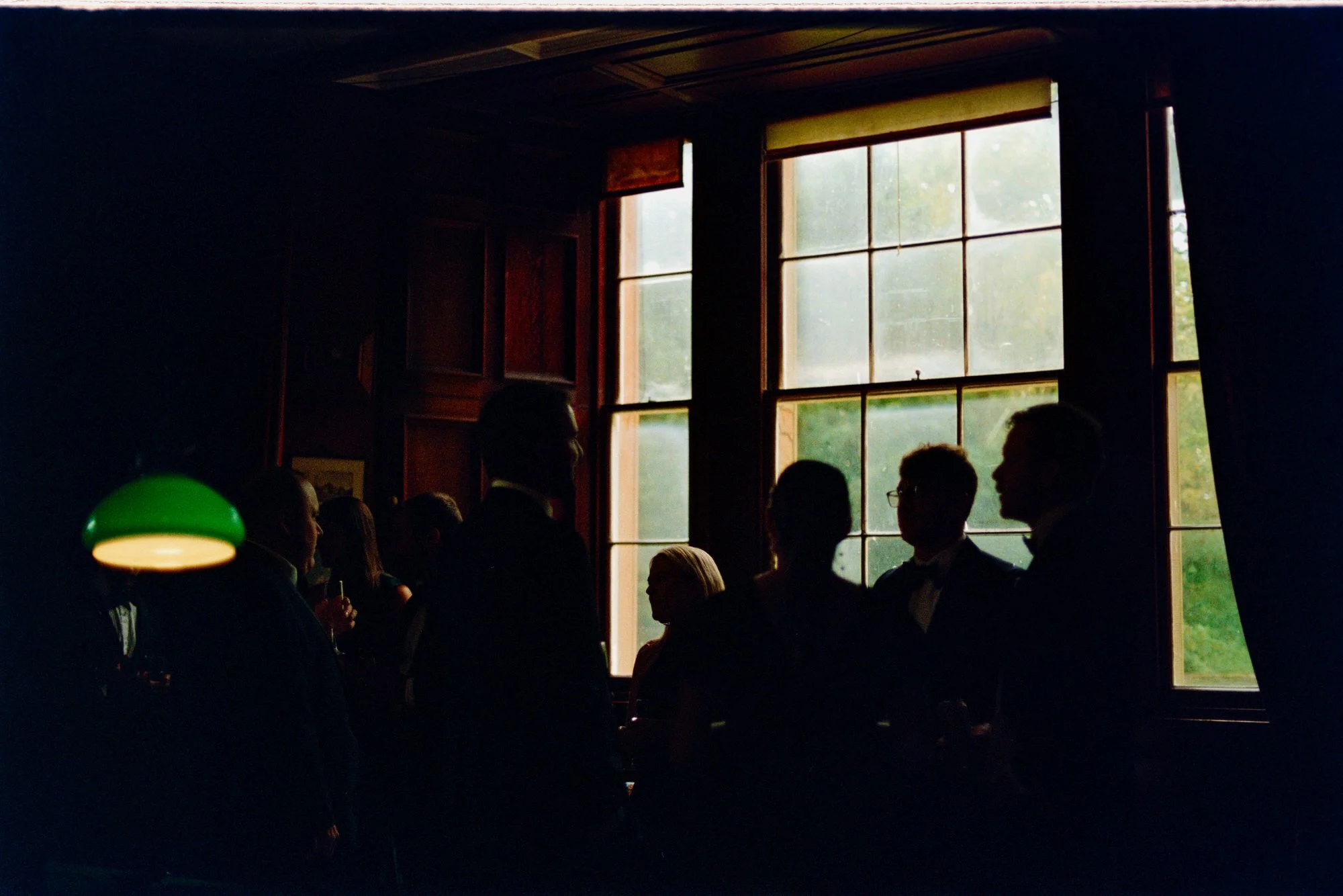 Guests gathered in silhouette beside a tall castle window and green reading lamp.