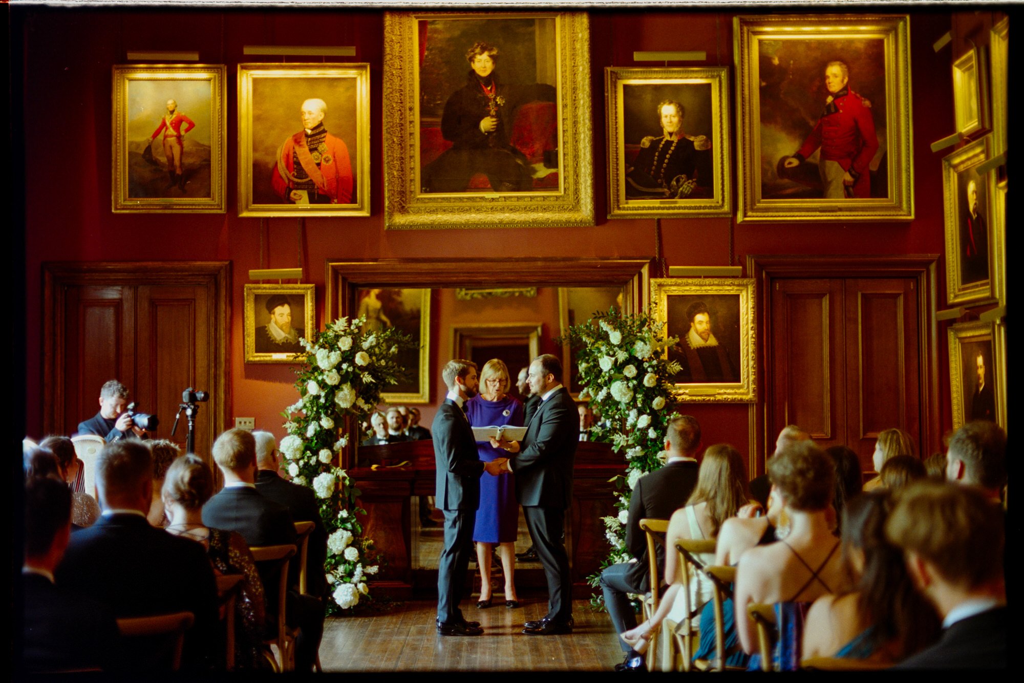 The grooms standing with their celebrant between floral arrangements in the State Dining Room.