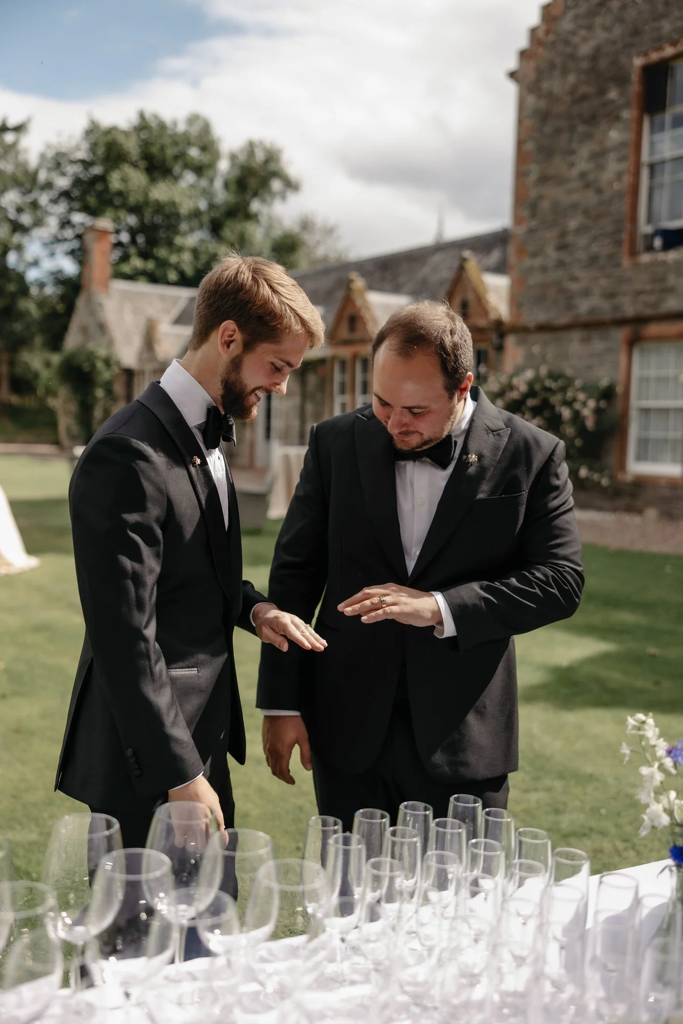 The newlyweds looking at their rings beside a table of glasses outdoors.