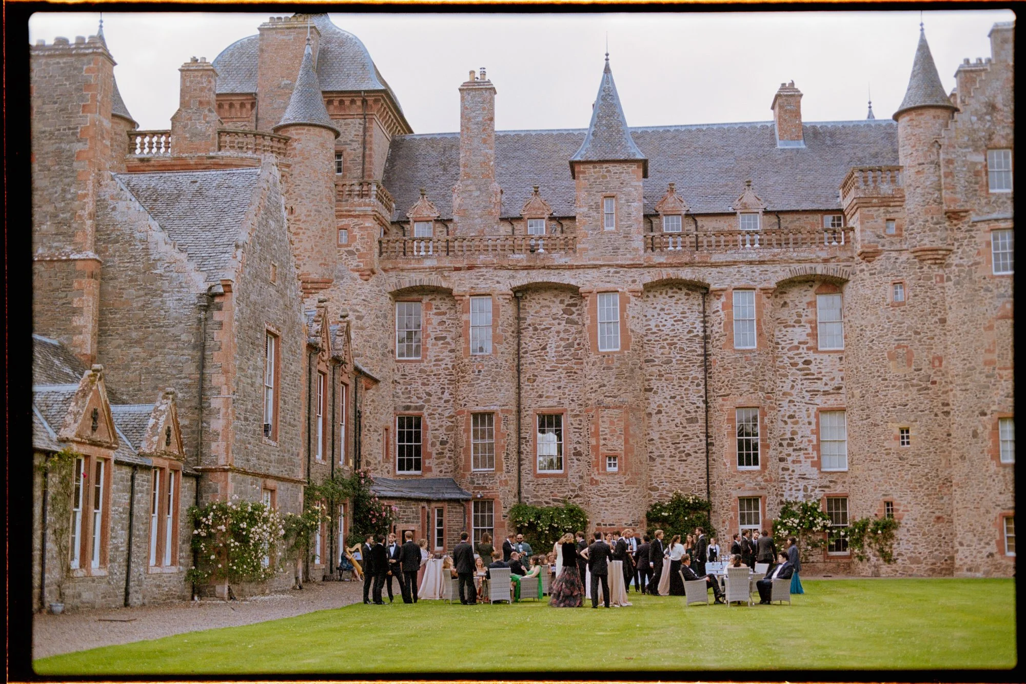 A wide film photograph of wedding guests gathered outside Thirlestane Castle.