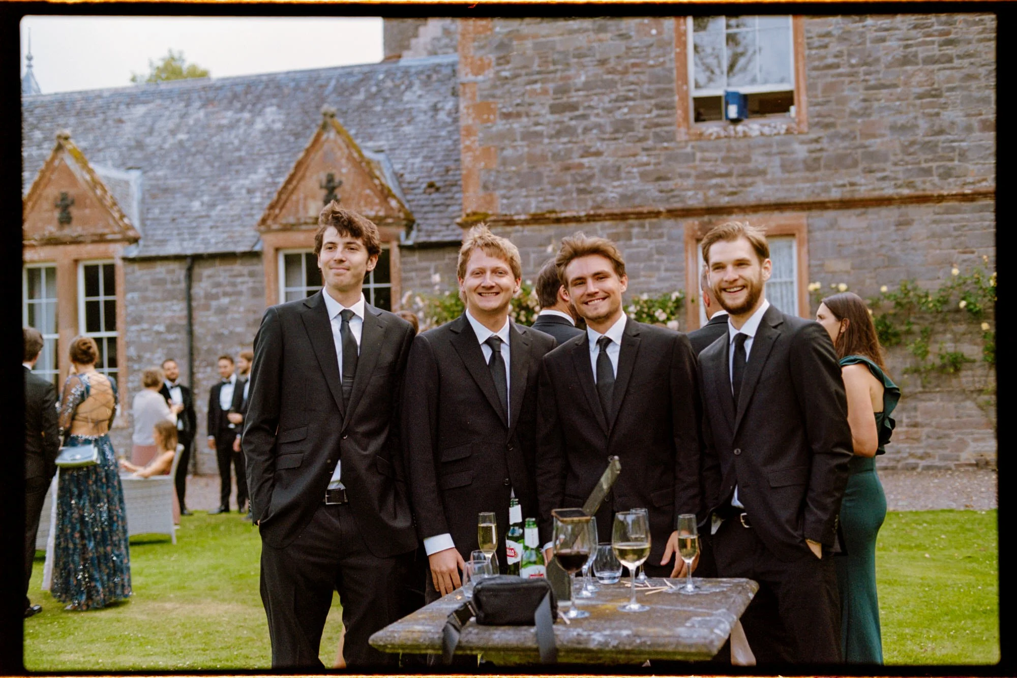 Four guests in dark suits posing beside a table of drinks outdoors.