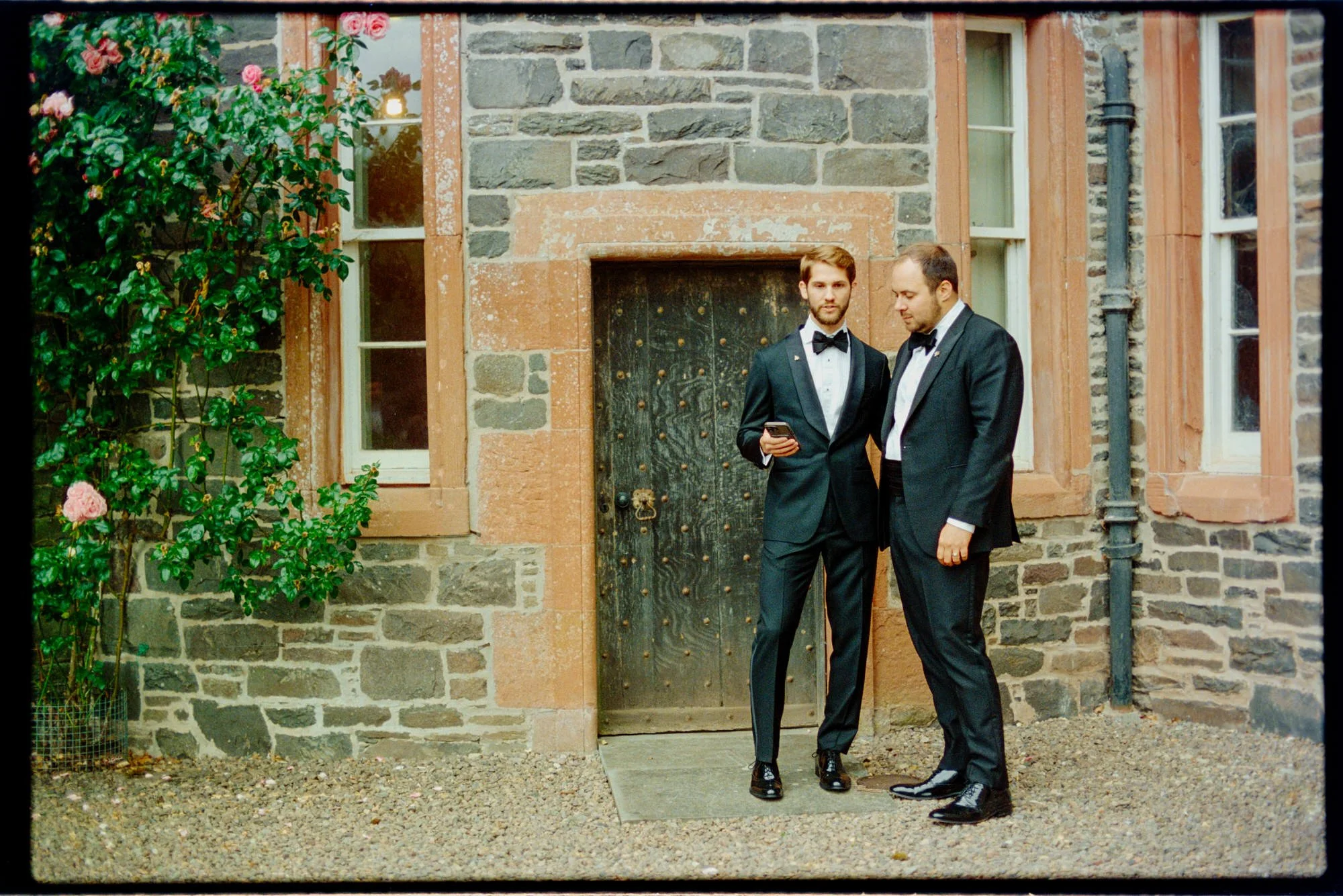 The newlyweds standing with drinks beside a stone doorway and climbing roses.