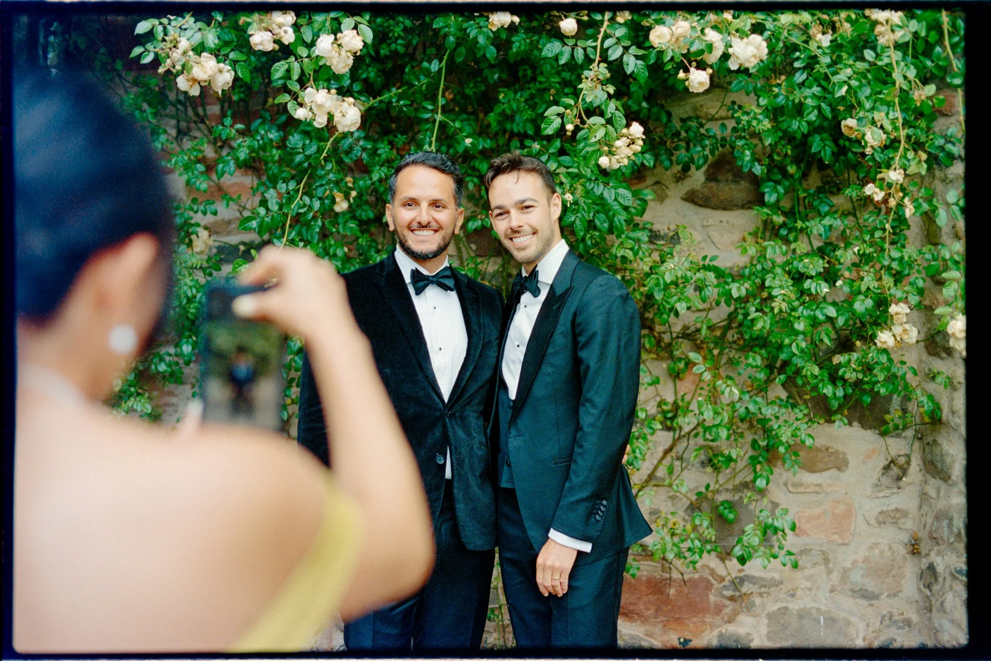 A guest taking a phone photograph of two guests beside pink roses.