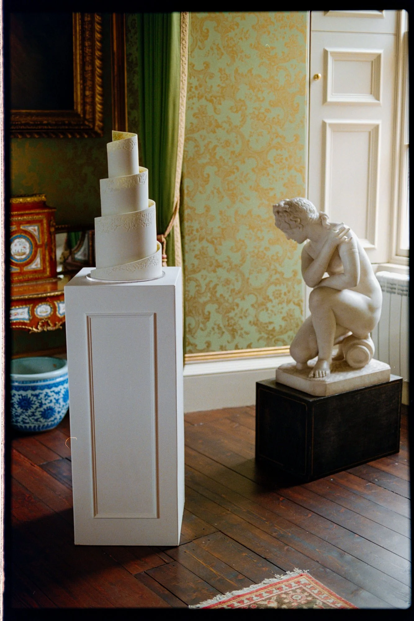 A tall sculptural wedding cake displayed beside a marble figure and castle window.