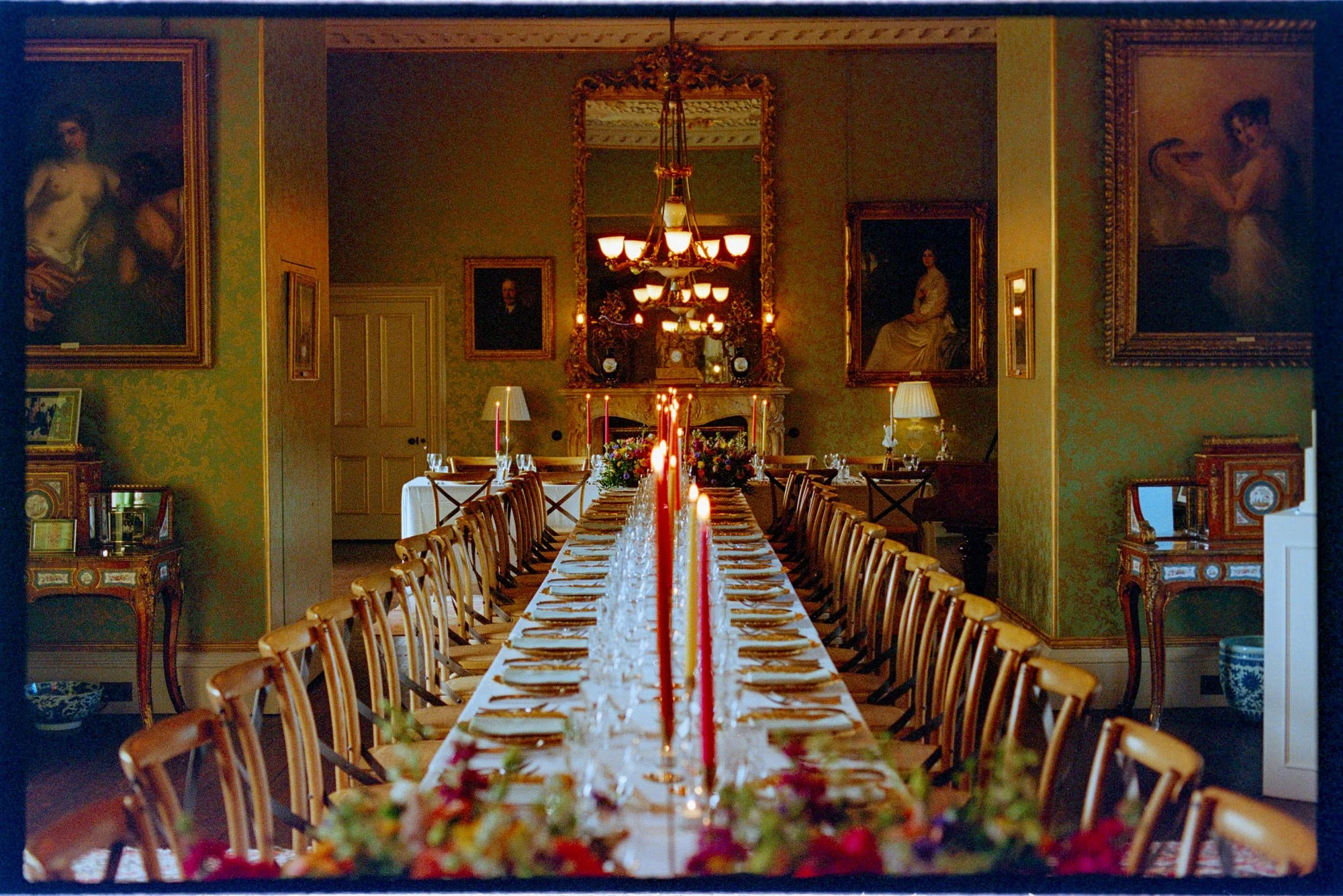 A long wedding dinner table set with flowers and red candles beneath a chandelier.