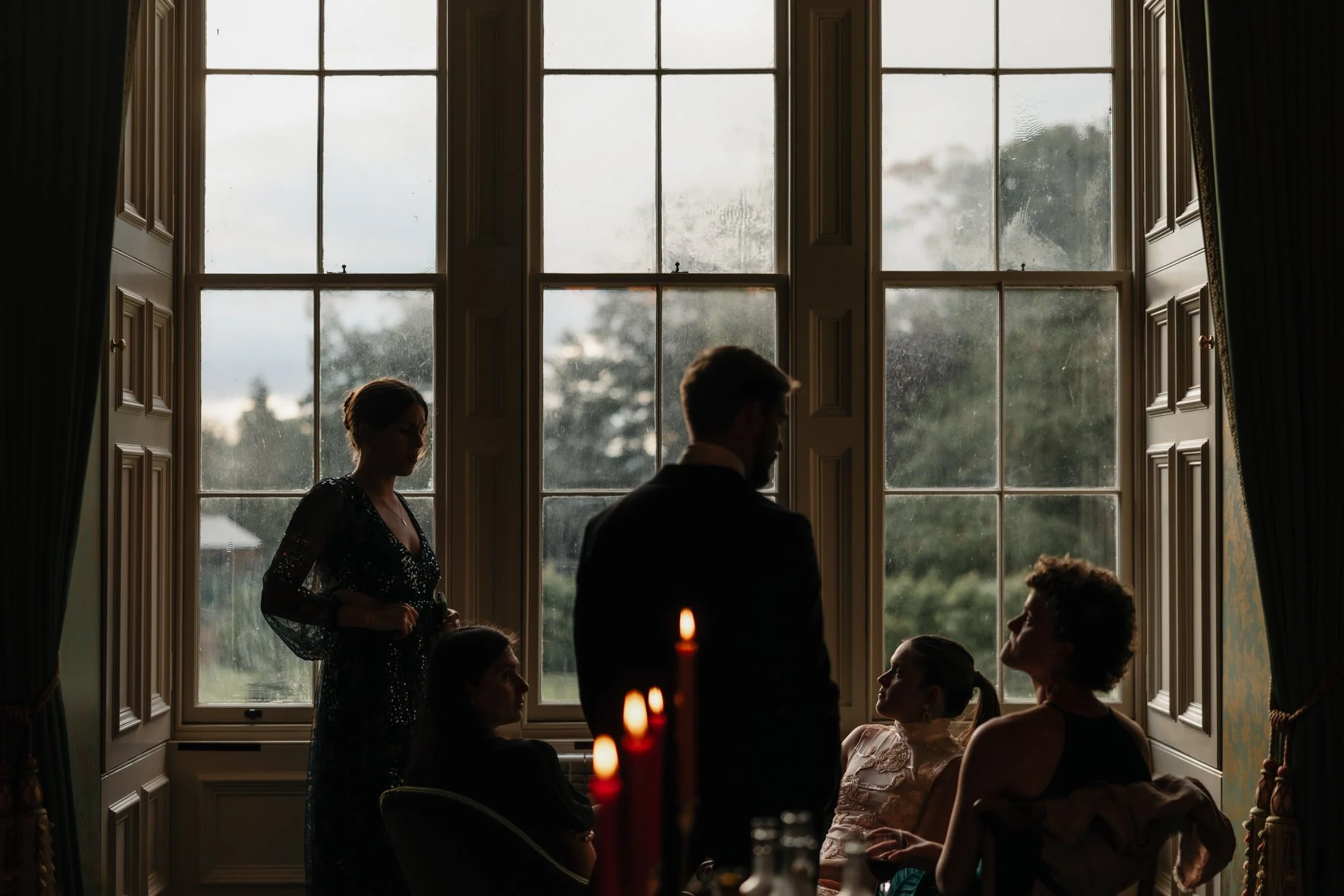 Guests talking beside a tall window as candles glow in the foreground.