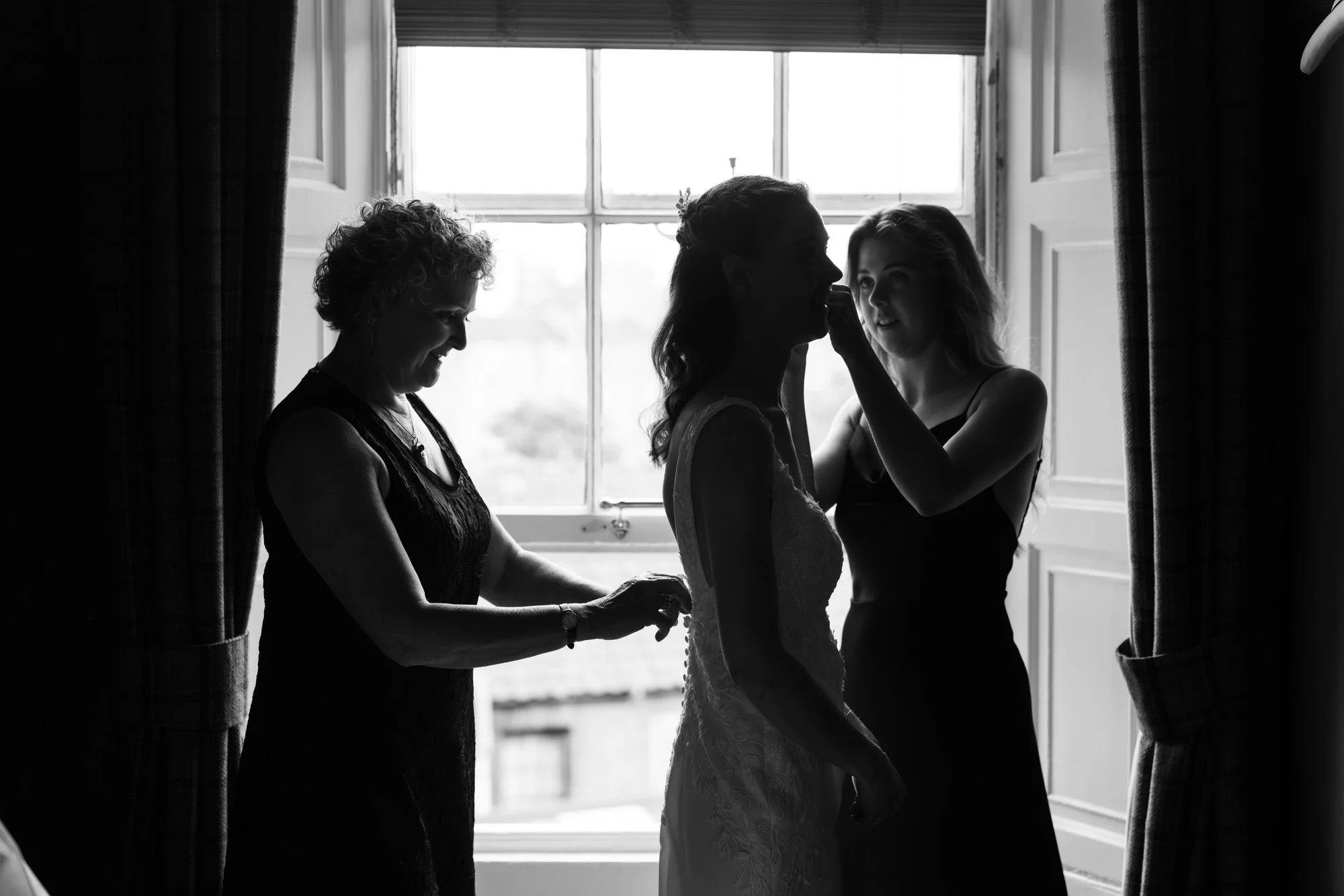 A bride helped into her dress beside the window
