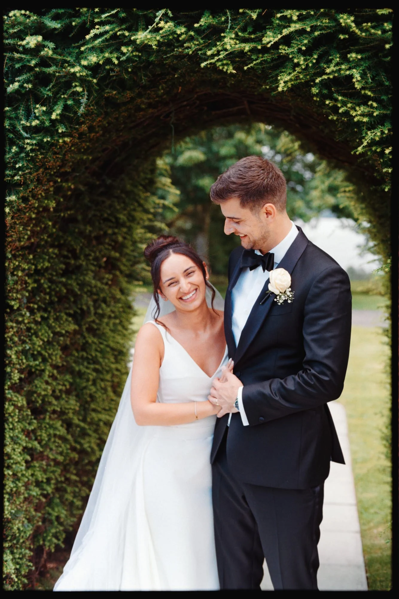 A newlywed couple standing under a green archway composed of hedge branches, smiling and looking at each other, with the bride in a white wedding dress and the groom in a black tuxedo with a boutonnière.