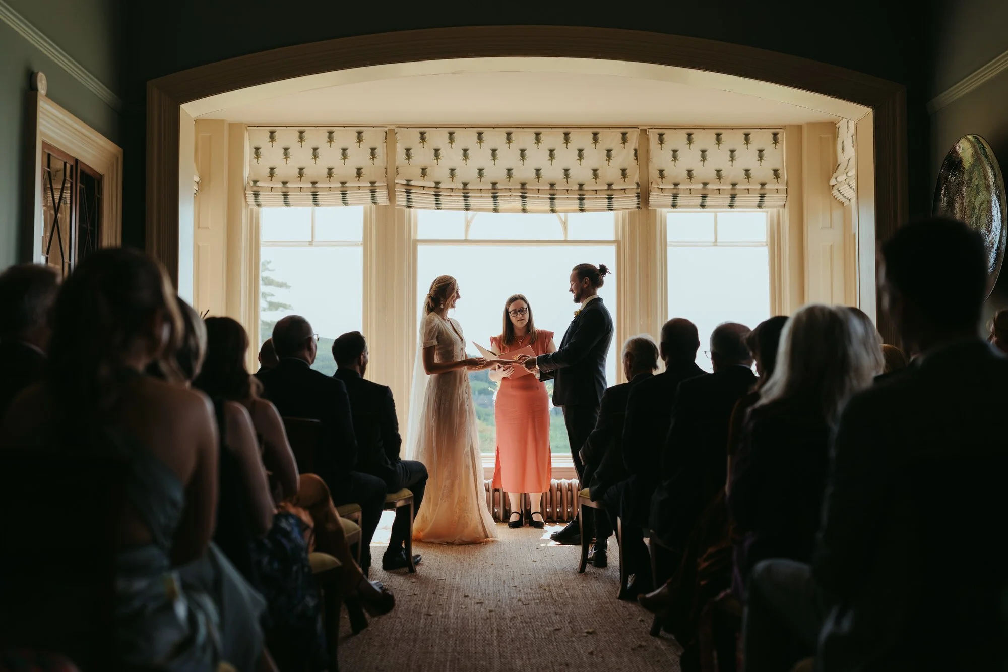 A wedding ceremony with a couple standing before an officiant, exchanging vows in front of a large window with bright natural light, surrounded by seated guests in an elegant indoor setting.