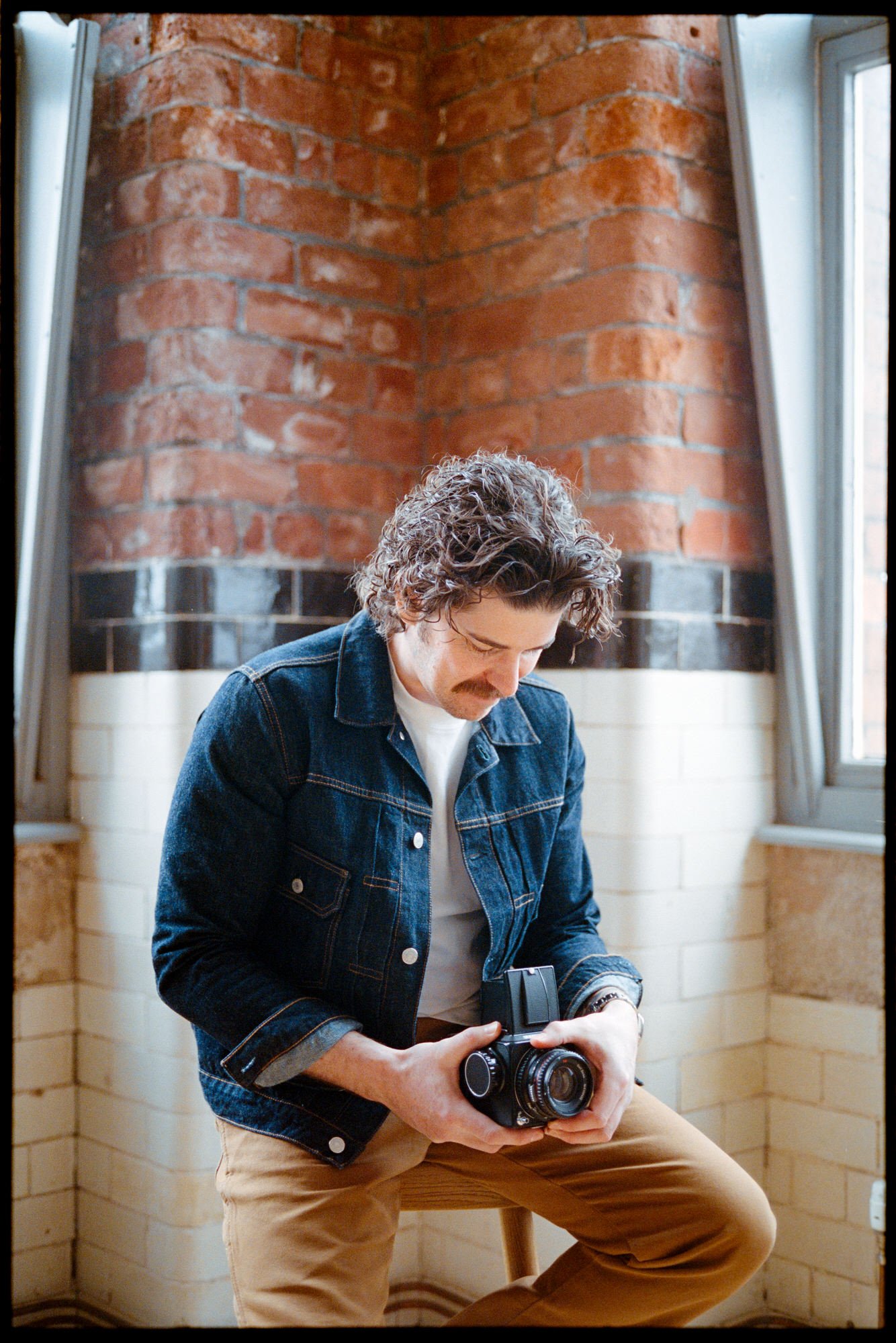 A man with curly hair and a mustache, wearing a denim jacket and khaki pants, holding a vintage camera while sitting on a wooden stool in a room with red brick walls and two windows. Photographer Steven William Weir, Edinburgh Scotland.