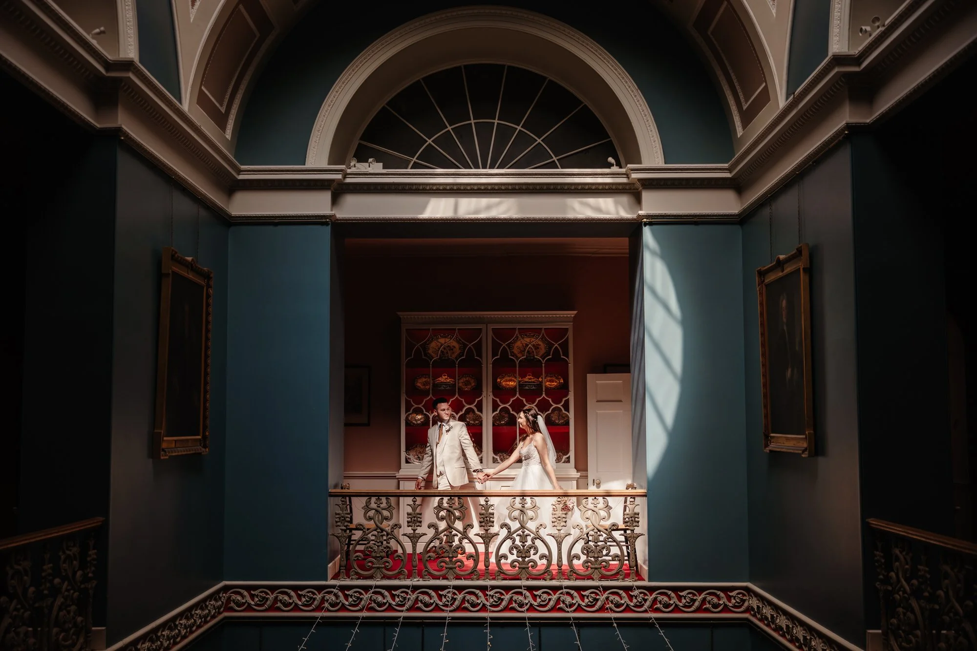 A bride and groom holding hands on a balcony in elegant surroundings with ornate decor, paintings, and blue walls. Photographed by Steven William Weir, Edinburgh Scotland.