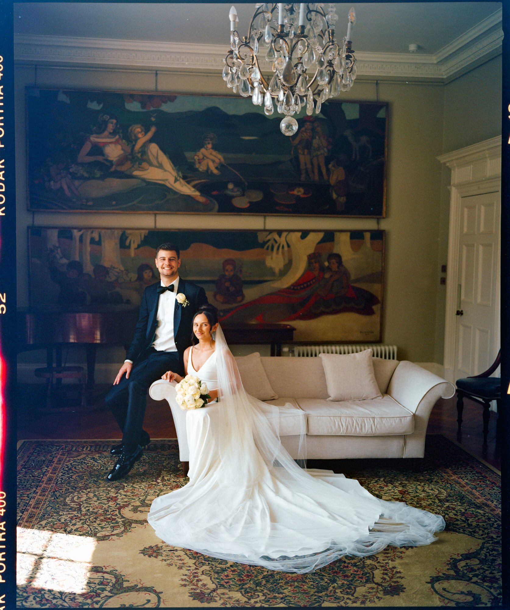 A newlywed couple in wedding attire posing in an elegant room with paintings, a chandelier, and a patterned rug. Photographed by Steven William Weir, Edinburgh Scotland.