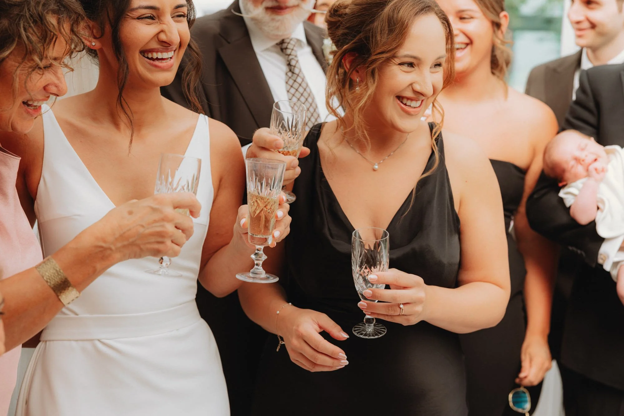 Group of people at a celebration, holding glasses of champagne, smiling and talking, with a baby in a person's arms in the background.