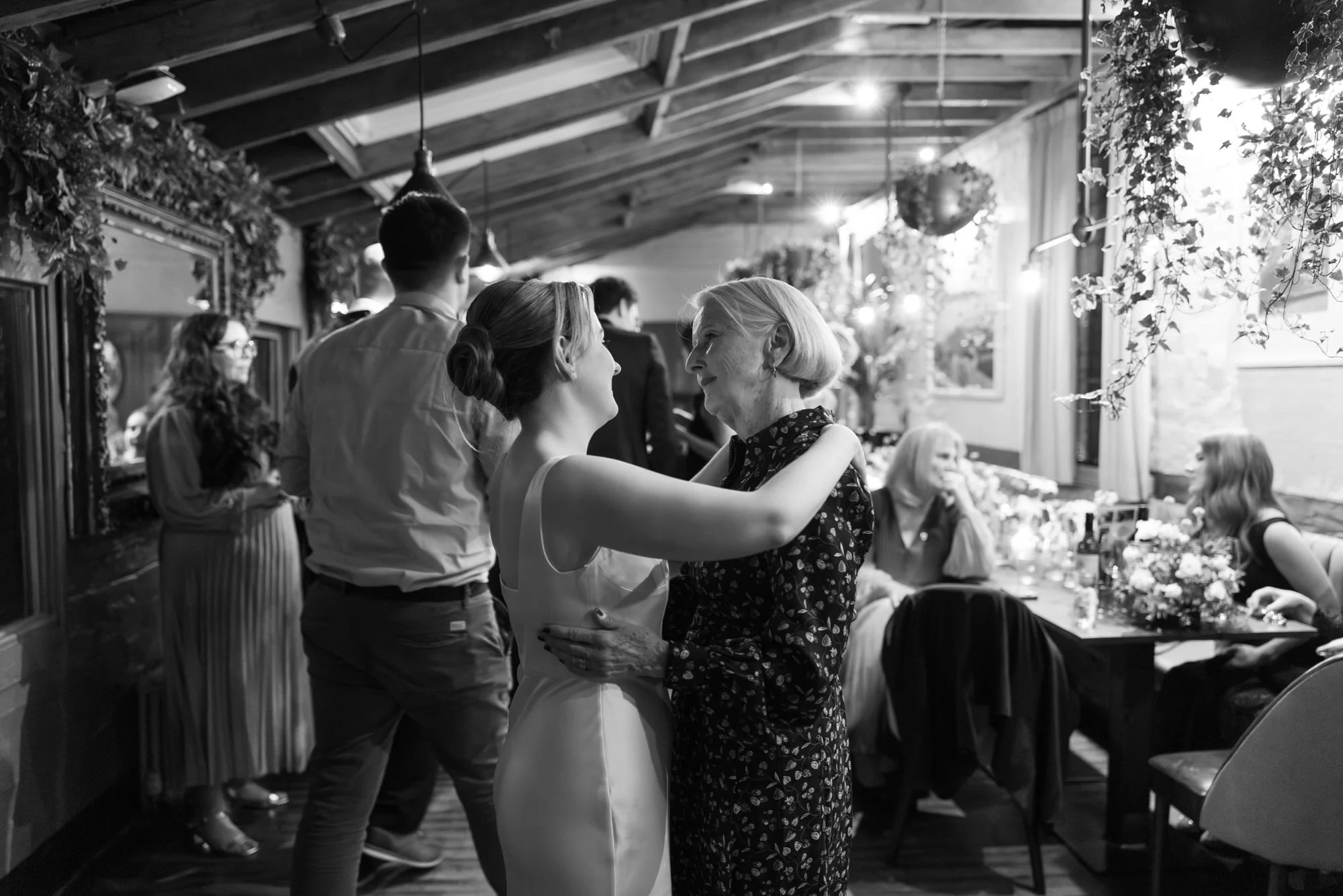 A bride and an elderly woman sharing a tender dance at a wedding reception in a decorated venue with hanging plants and flower arrangements, while guests sit and stand around tables in the background.