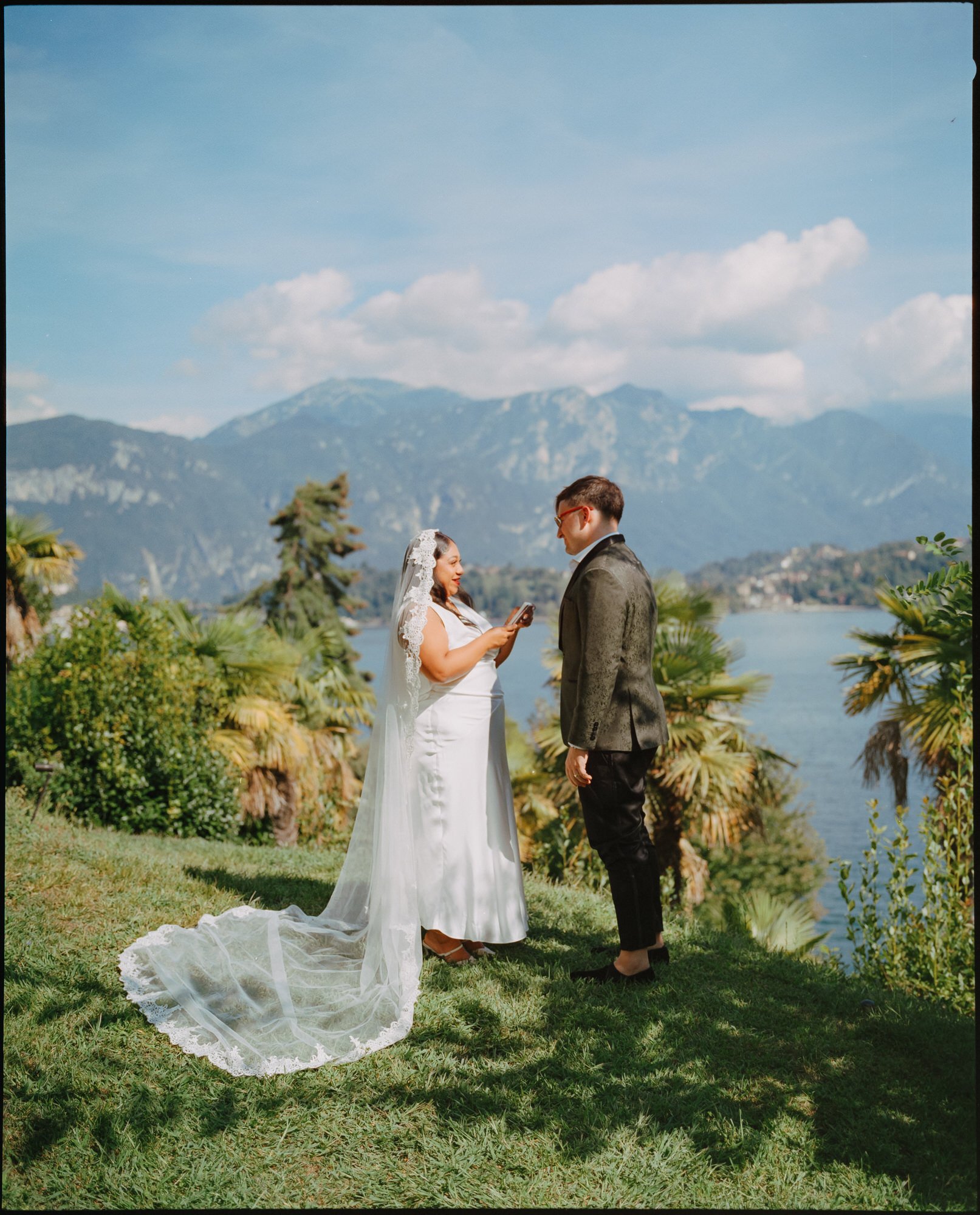 A bride and groom standing outdoors on green grass, facing each other with a lake and mountains in the background, during a wedding ceremony. Photographed by Steven William Weir, Edinburgh Scotland.