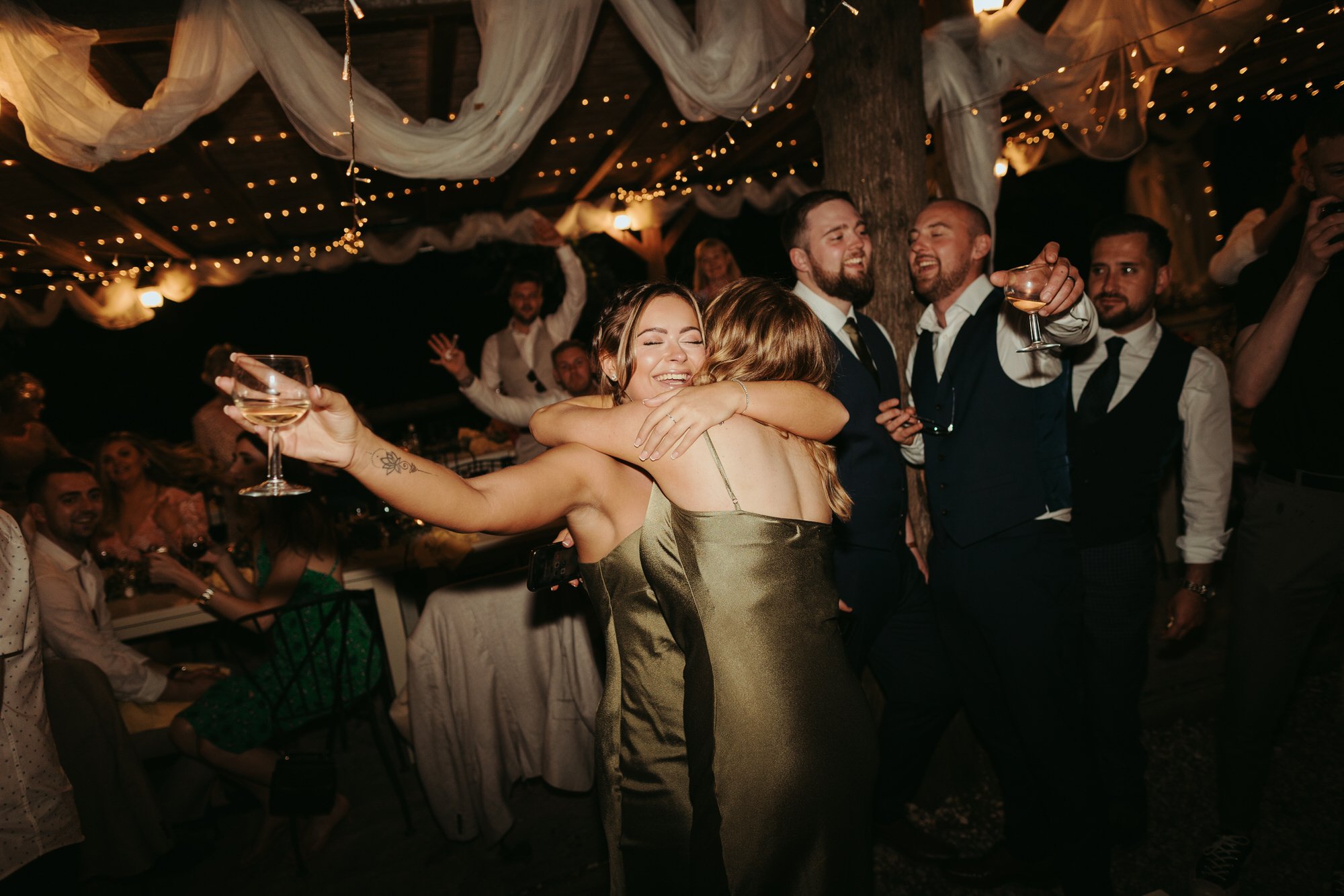 People celebrating at a wedding reception, hugging, smiling, and holding drinks under string lights and decorated fabric.