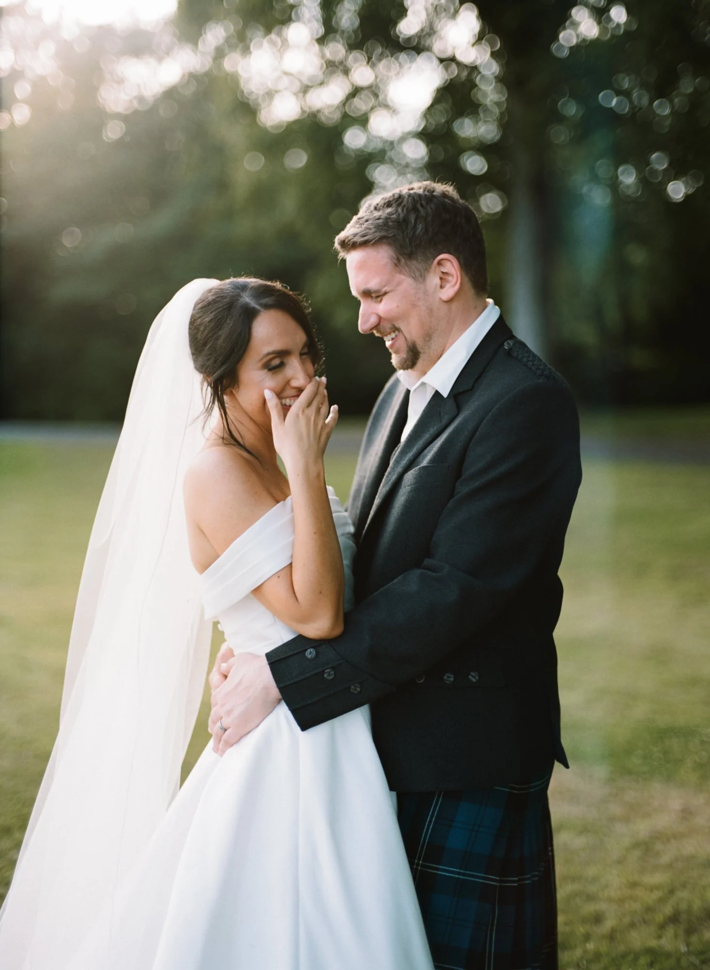 A bride and groom share a joyful moment outdoors during sunset, embracing and laughing together, with a blurred natural background. Photographed by Steven William Weir, Edinburgh Scotland.