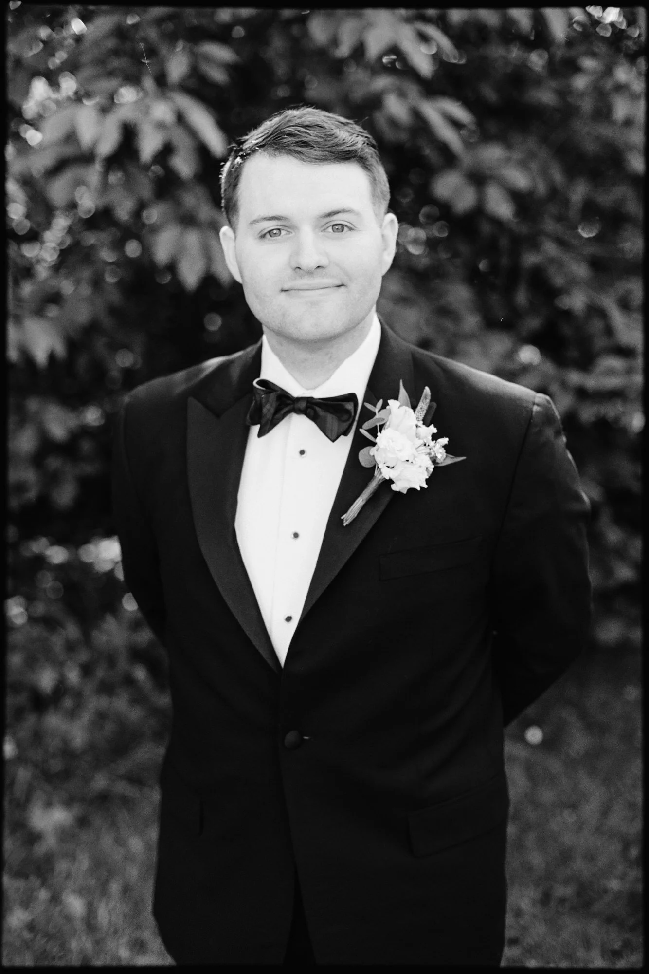 Black and white photo of a man in formal tuxedo with a boutonniere, standing outdoors.
