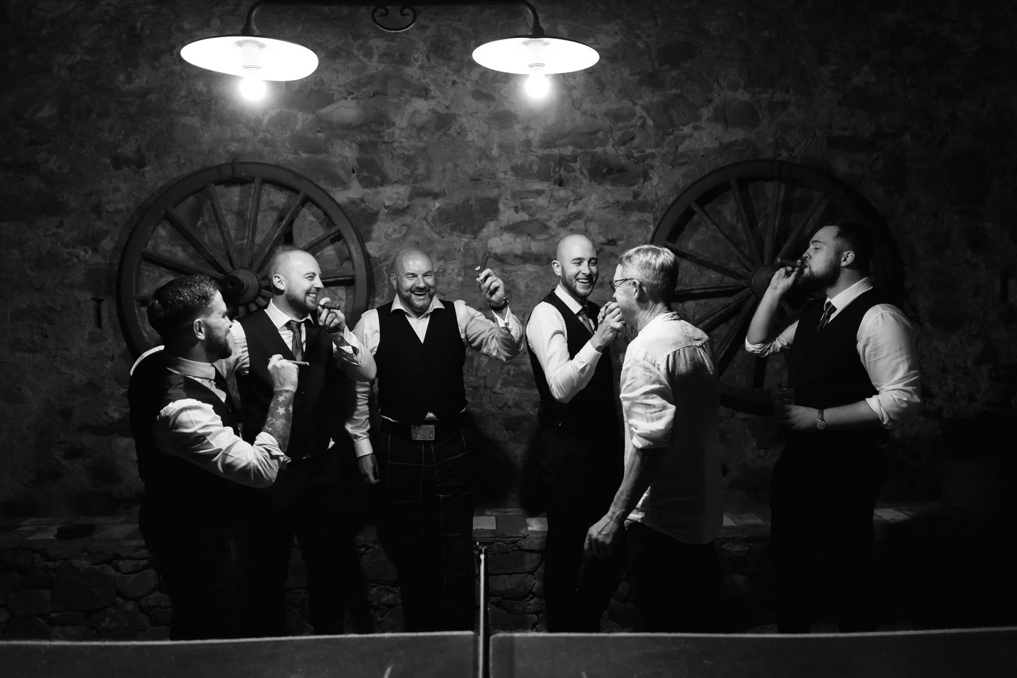 Black and white photo of six men standing in a line in a rustic setting with stone walls and wagon wheels, appear to be celebrating or socializing, some holding cigars and drinks, smiling and laughing.