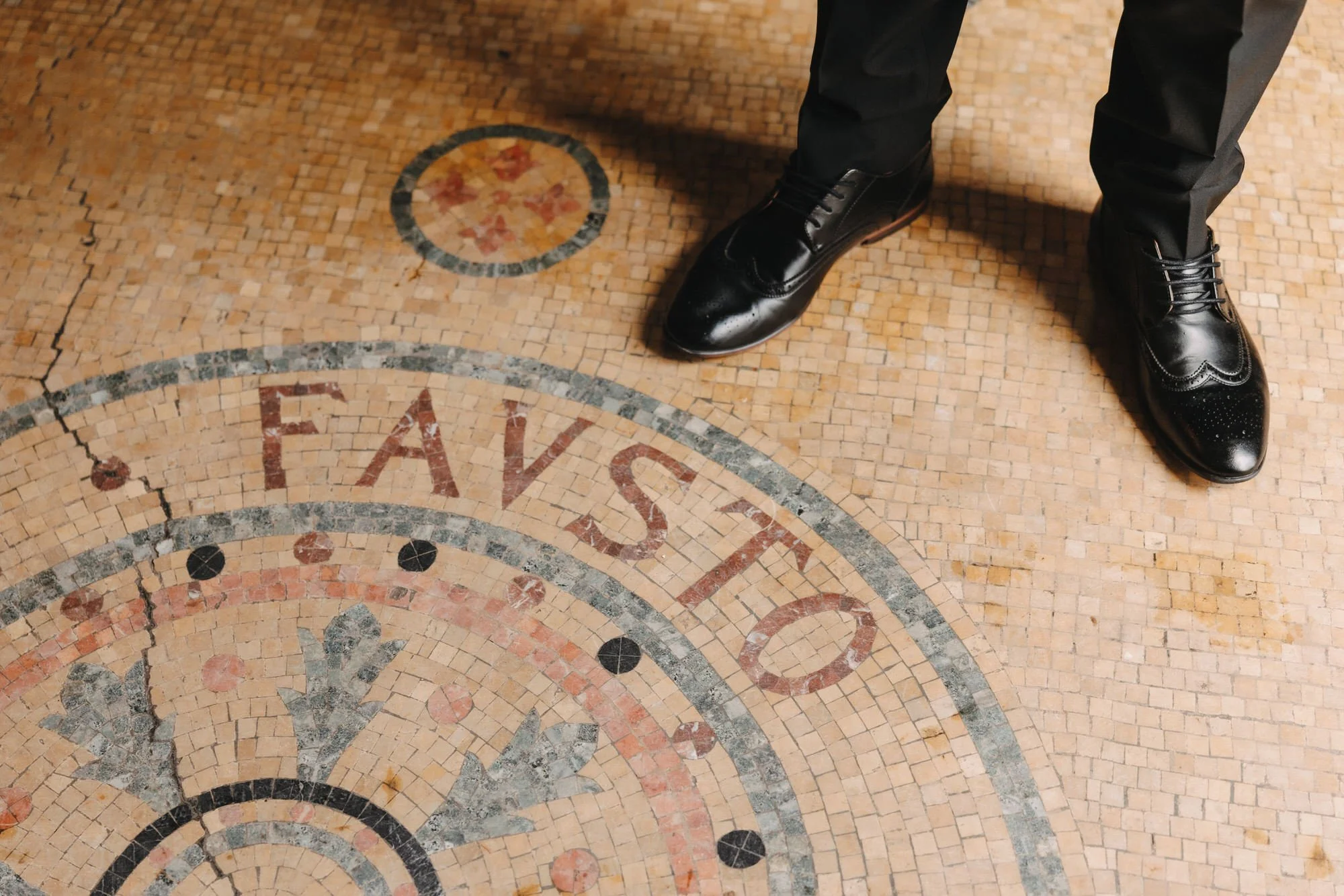 Person standing on a mosaic floor with the word 'FASTER' written on it, wearing black dress shoes and black pants.