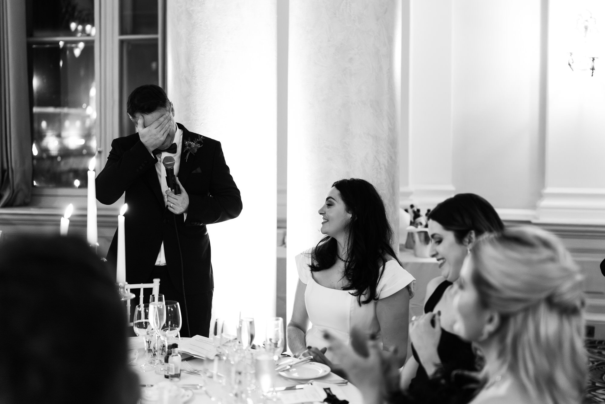 A man in a suit giving a speech at a wedding reception, covering his face with one hand, while a woman in a white dress listens and smiles at the table with other women.