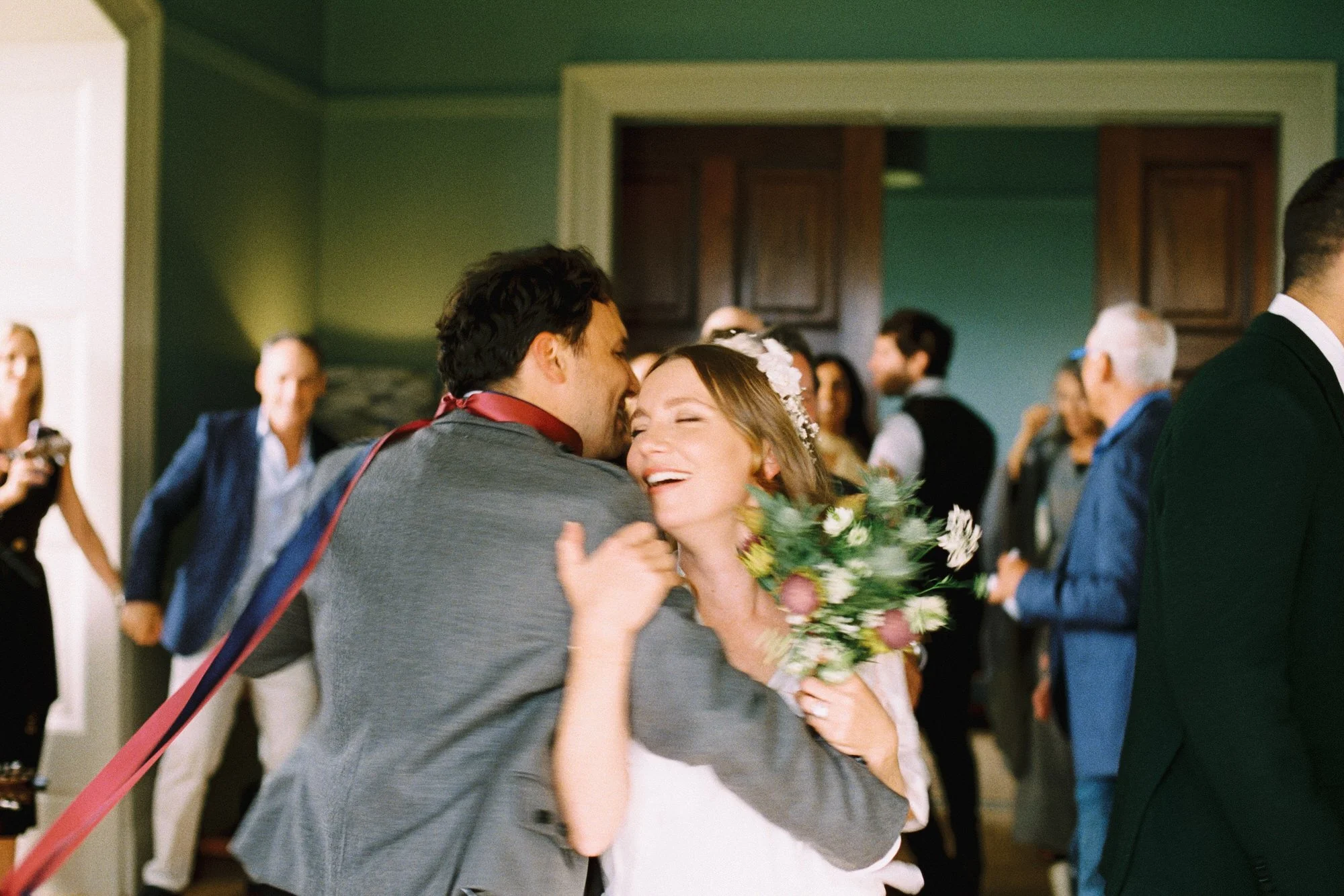 A couple embracing and smiling at each other at a wedding reception. The woman is holding a bouquet of flowers and the man is wearing a gray suit. They are surrounded by guests.