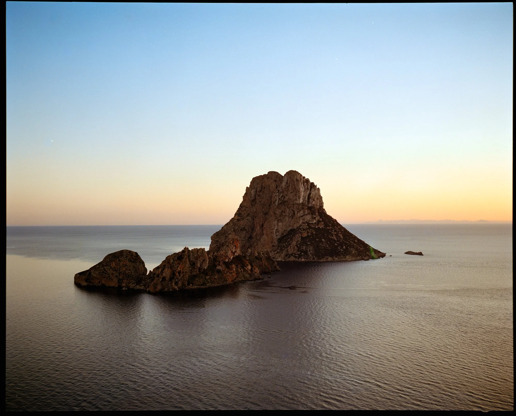 A large rocky island surrounded by calm ocean waters at sunset or sunrise, with a clear sky and distant landforms on the horizon.