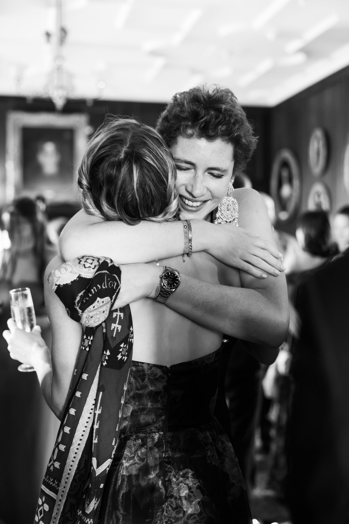 Two women hugging at a social event, one wearing a strapless dress and the other with her back to the camera, holding a glass of champagne, with other people in the background. Photographed by Steven William Weir, Edinburgh Scotland.