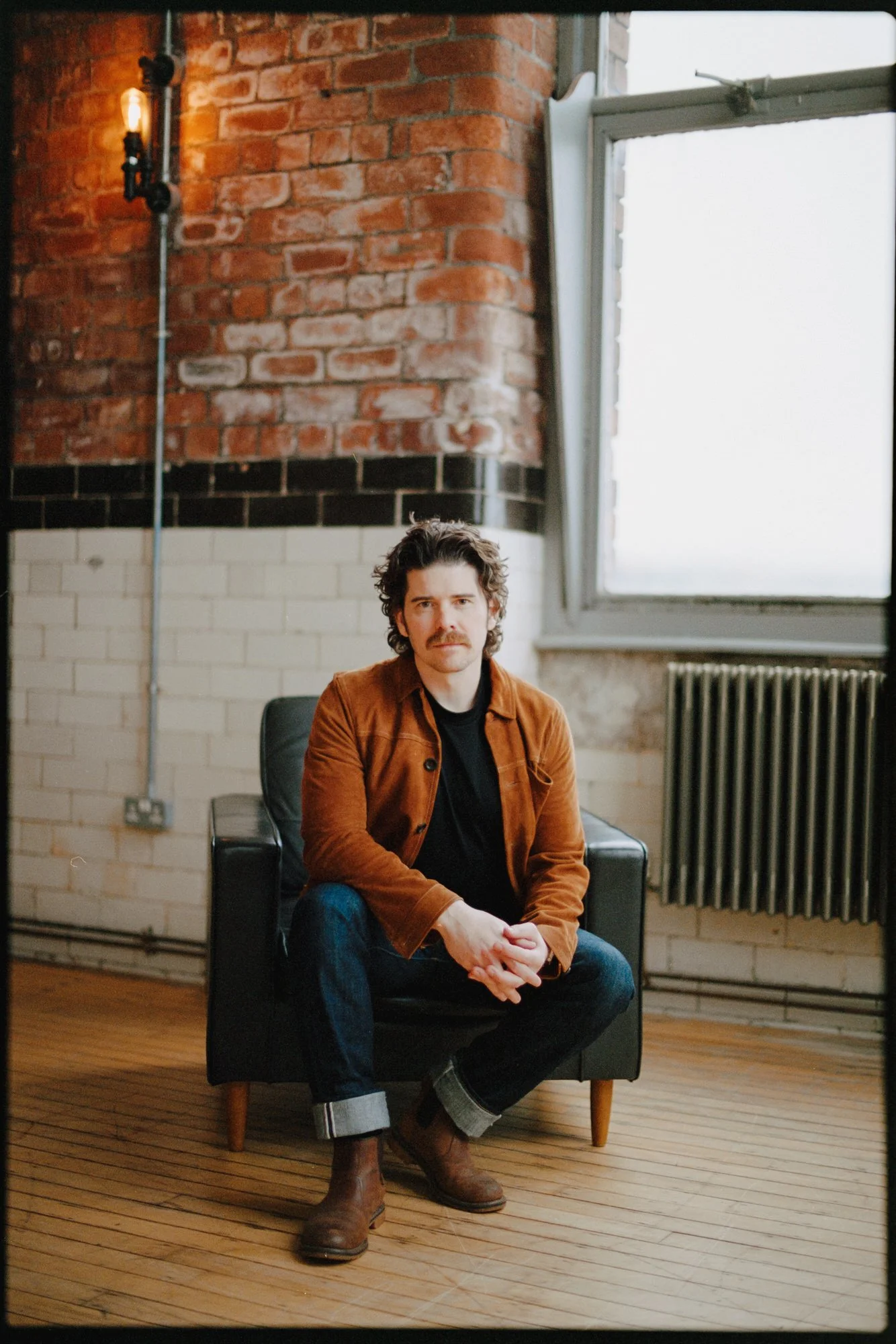 A man with curly dark hair, a mustache, wearing a brown jacket, black shirt, cuffed jeans, and brown boots, sitting on a black armchair in a room with exposed brick and white painted brick walls, wooden floor, and a large open window.