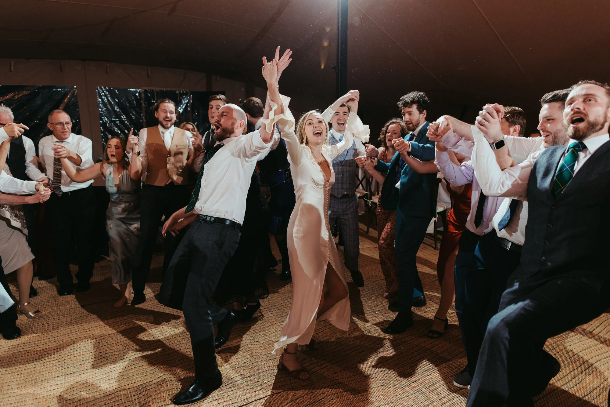 Group of people dancing and celebrating at a wedding reception inside a large tent.