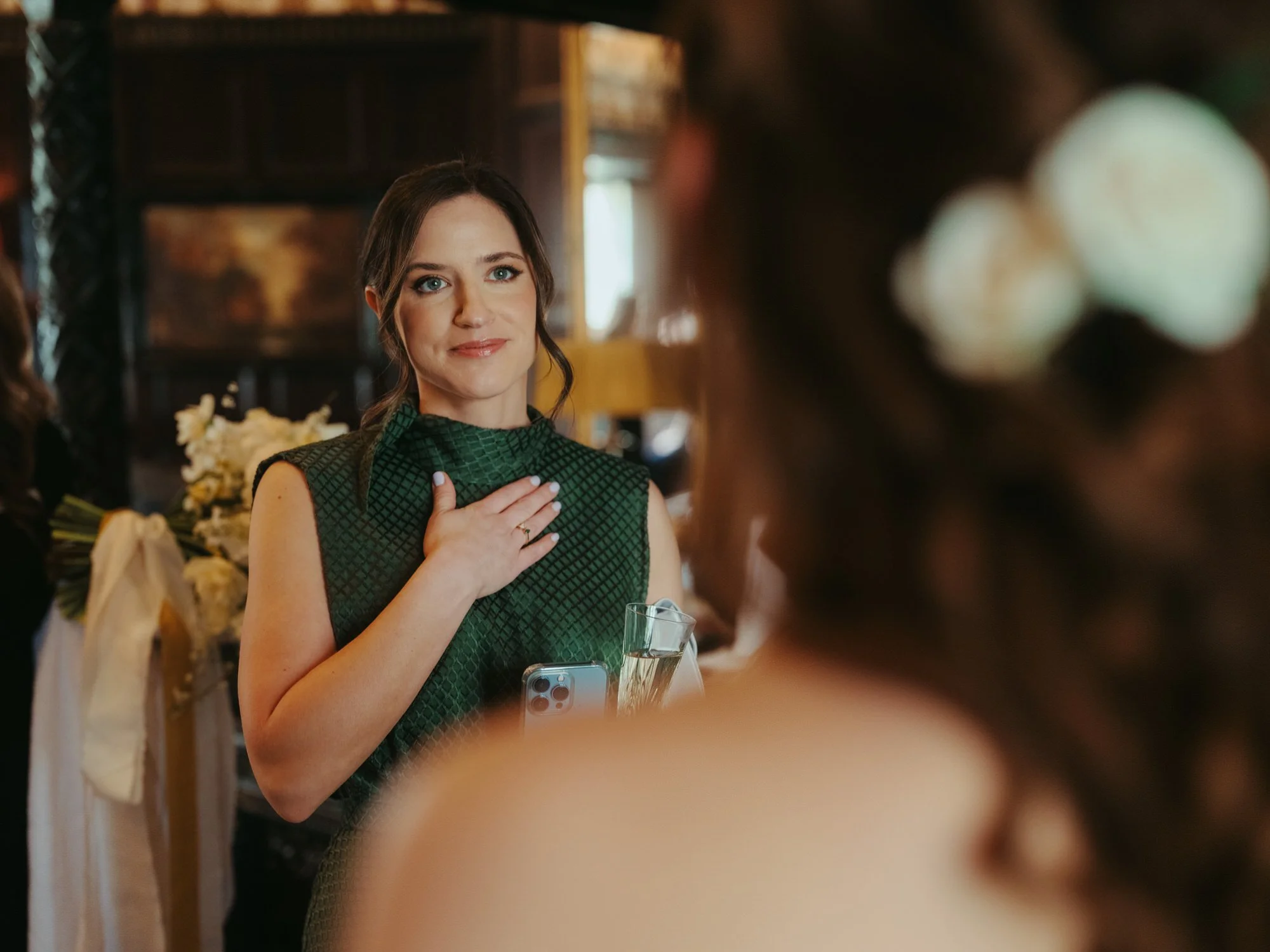 A woman in a green dress with diamond pattern, holding her hand over her chest and smiling, at a wedding reception. She has dark hair and is standing near another woman with white flowers in her hair.
